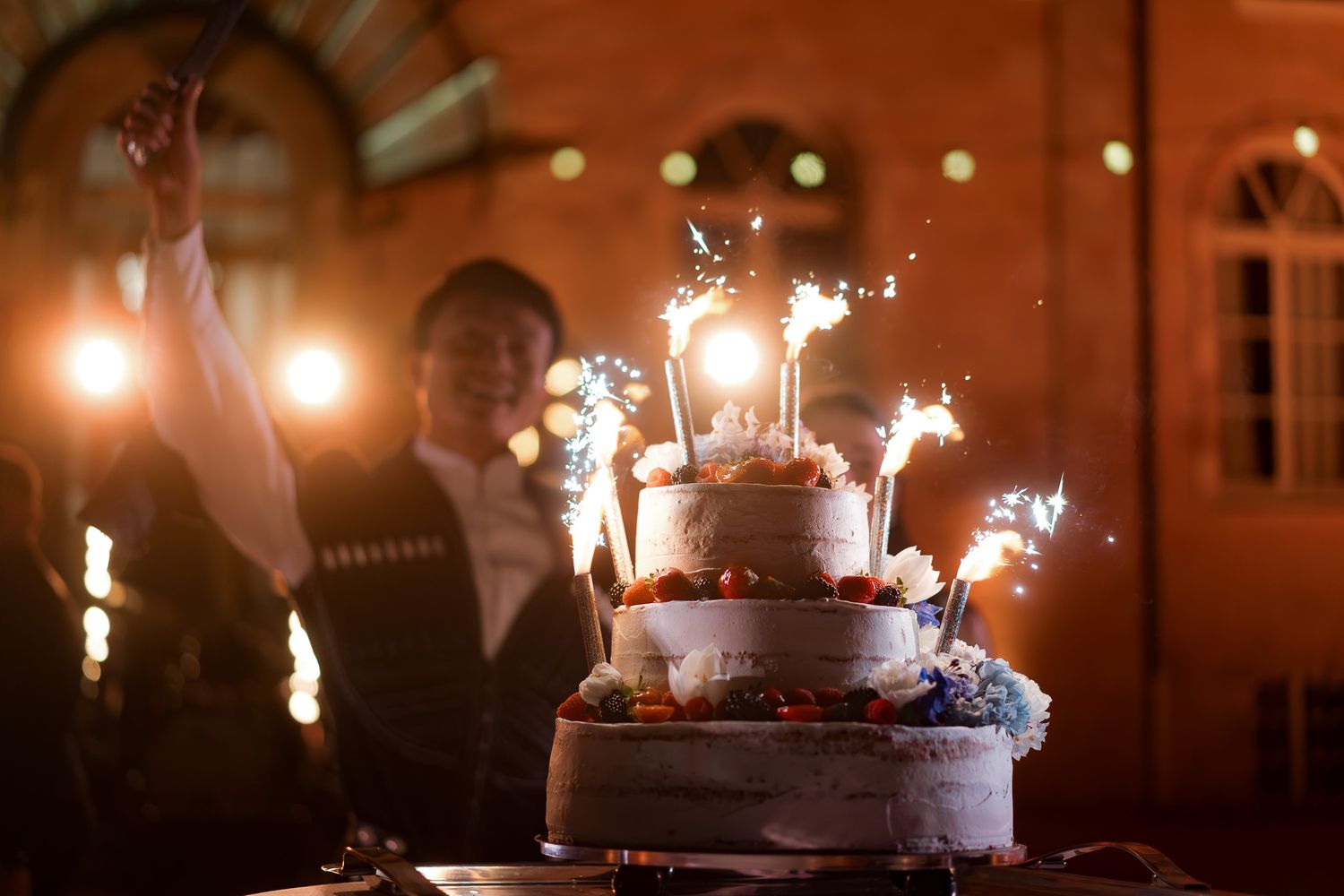 Three-tier wedding cake with sparklers glowing at night, guests celebrating in the background.