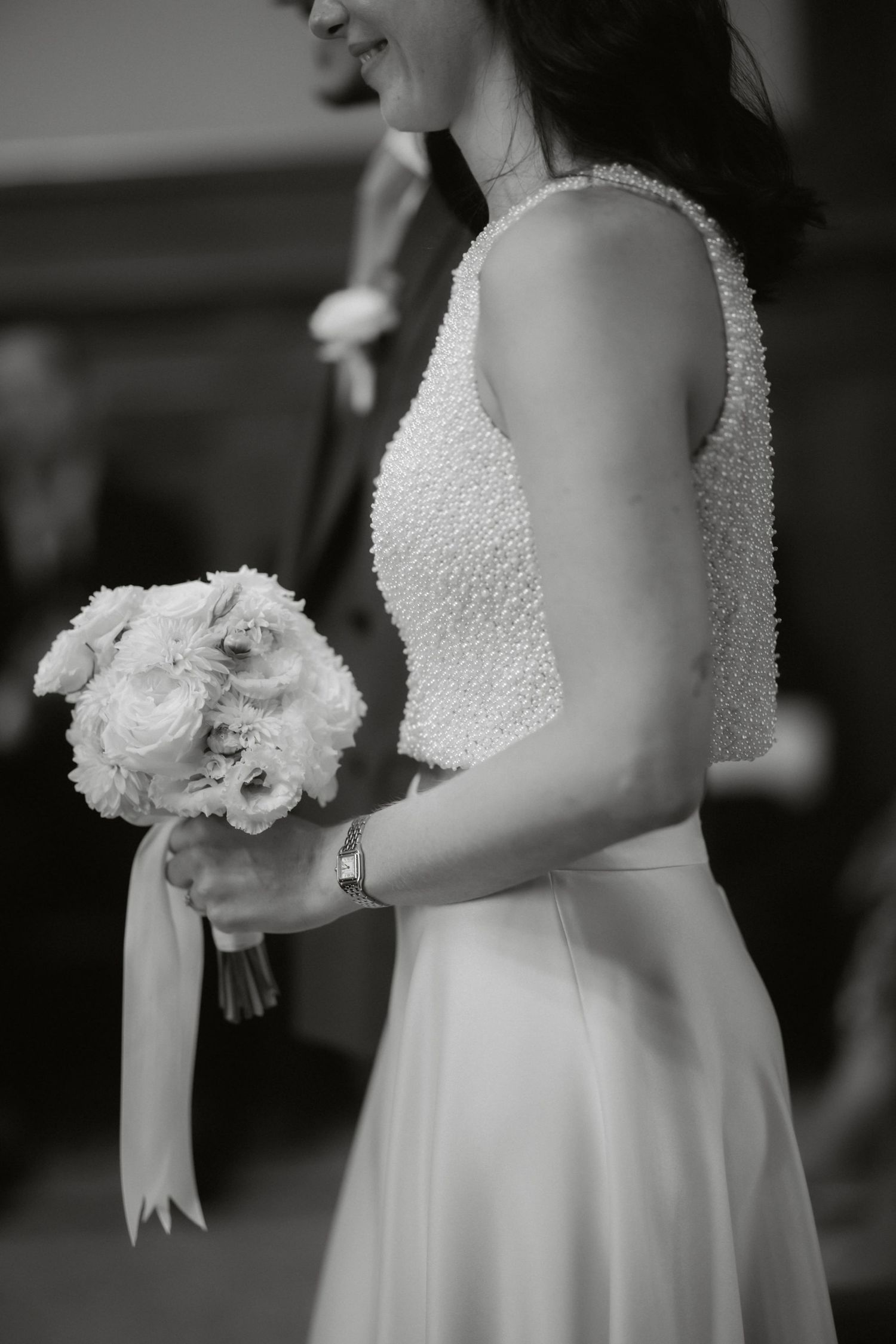 Black and white photo of a bride in a pearl-embellished gown holding a white rose bouquet with ribbon.