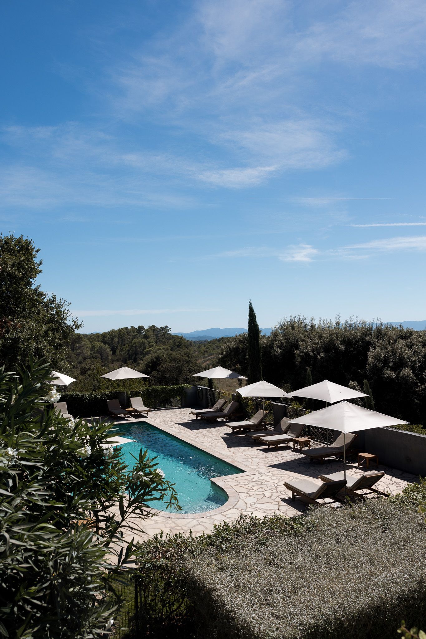 Luxury outdoor pool with lounge chairs and white umbrellas surrounded by lush greenery under a clear blue sky.