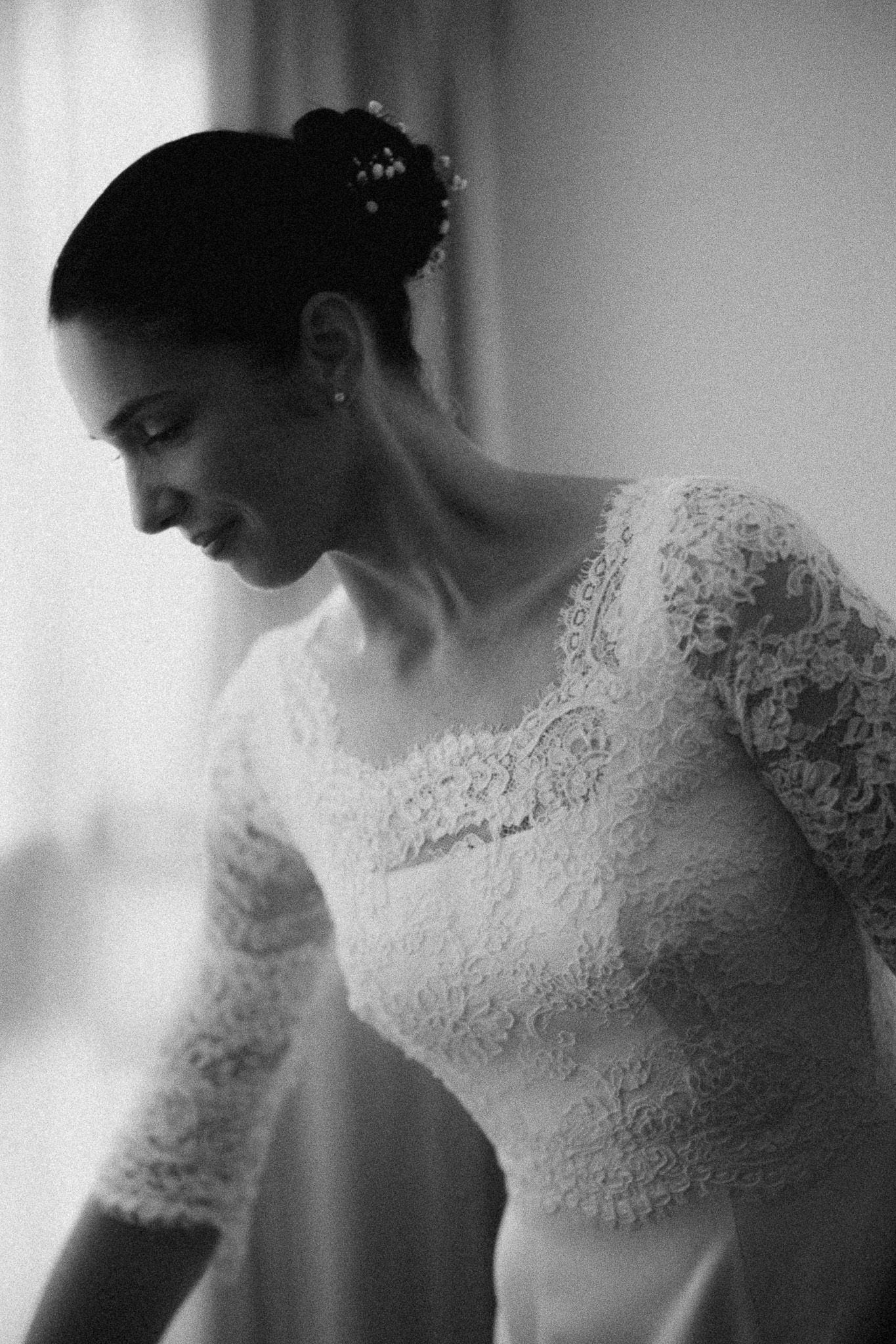 Black and white photo of a bride in lace wedding dress with elegant updo, smiling softly near a window.