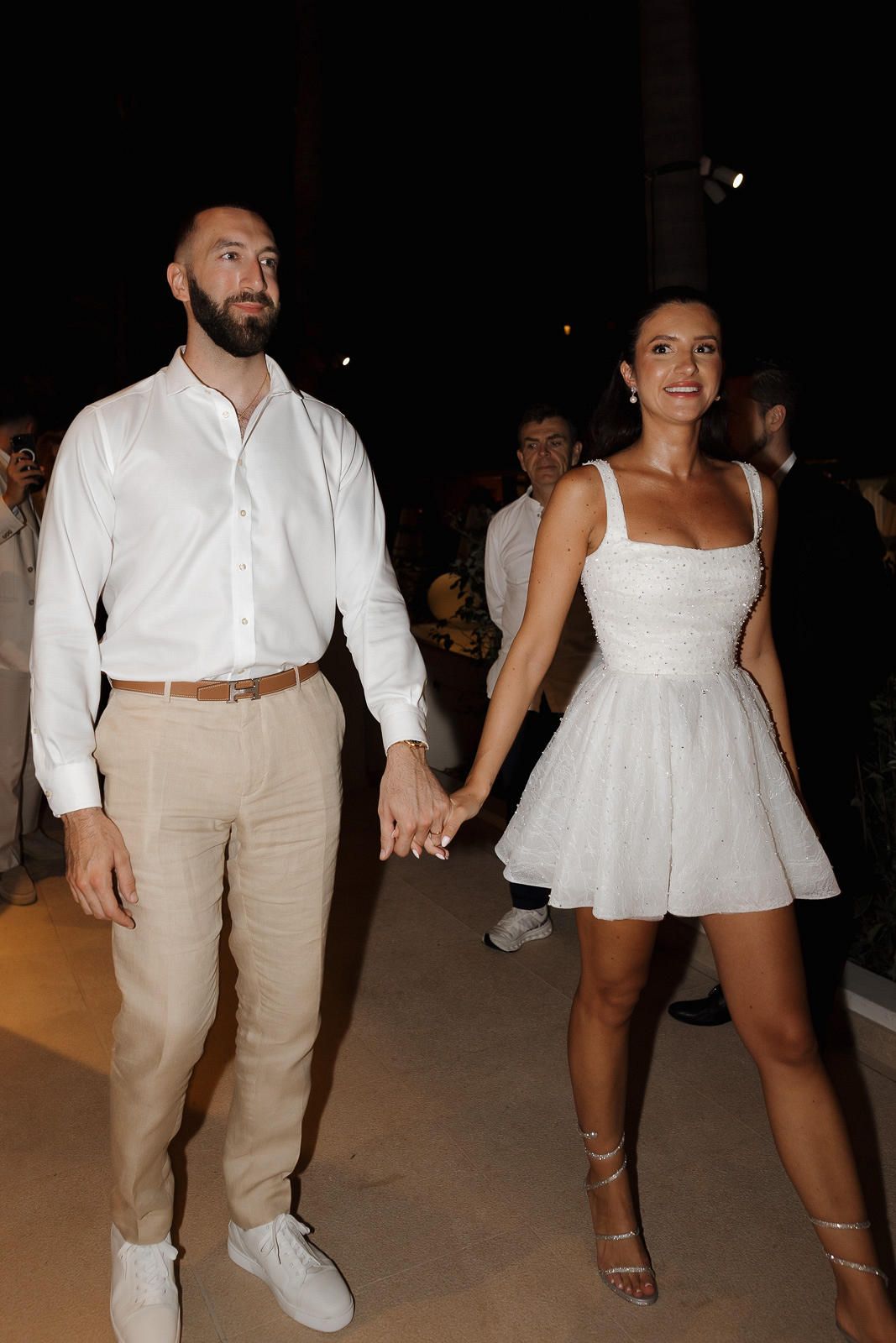 A couple holds hands at an evening event, with the man in white shirt and khakis and woman in a white mini dress.