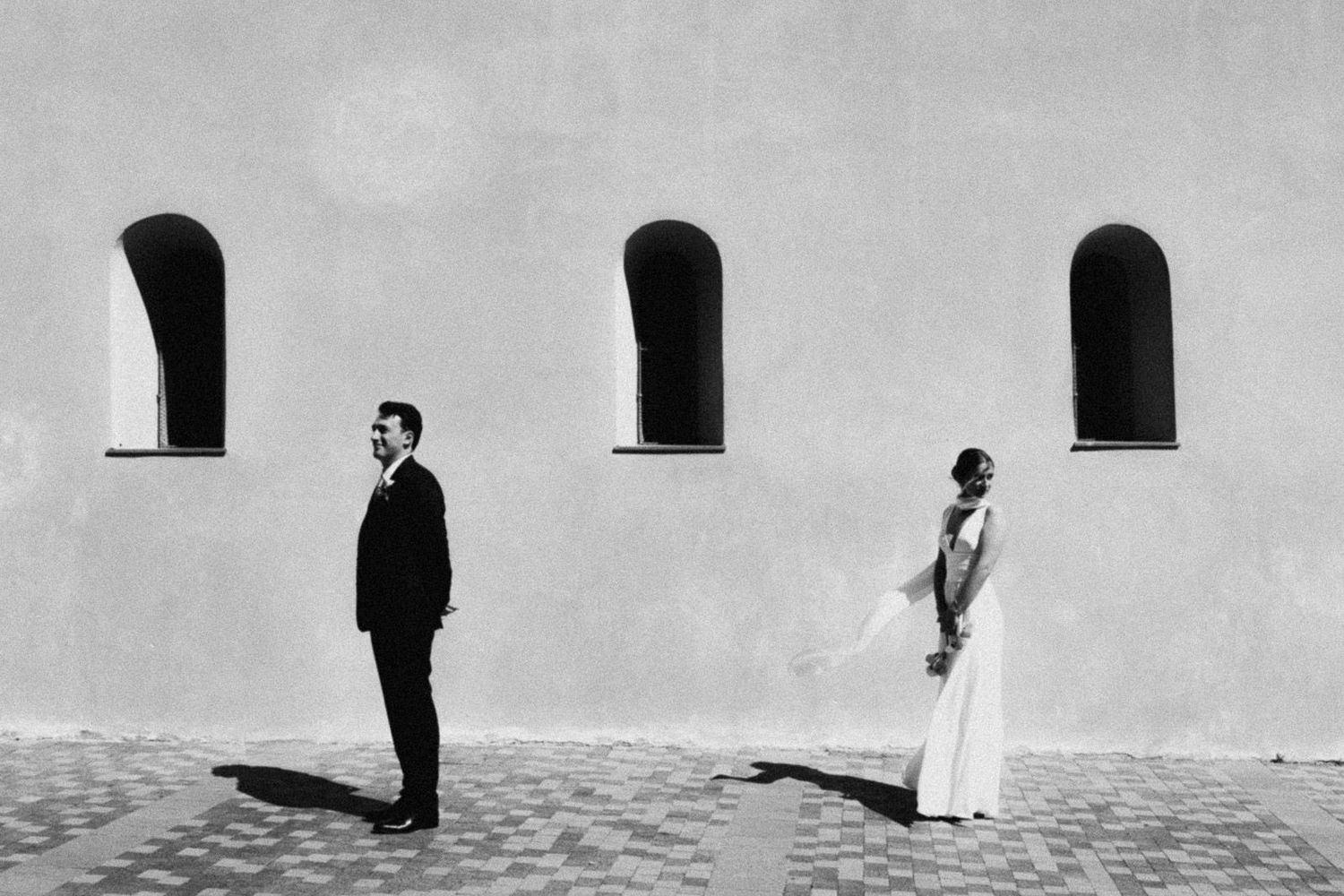 A minimalist black and white wedding photo with a couple standing apart against a white wall with three arched windows.