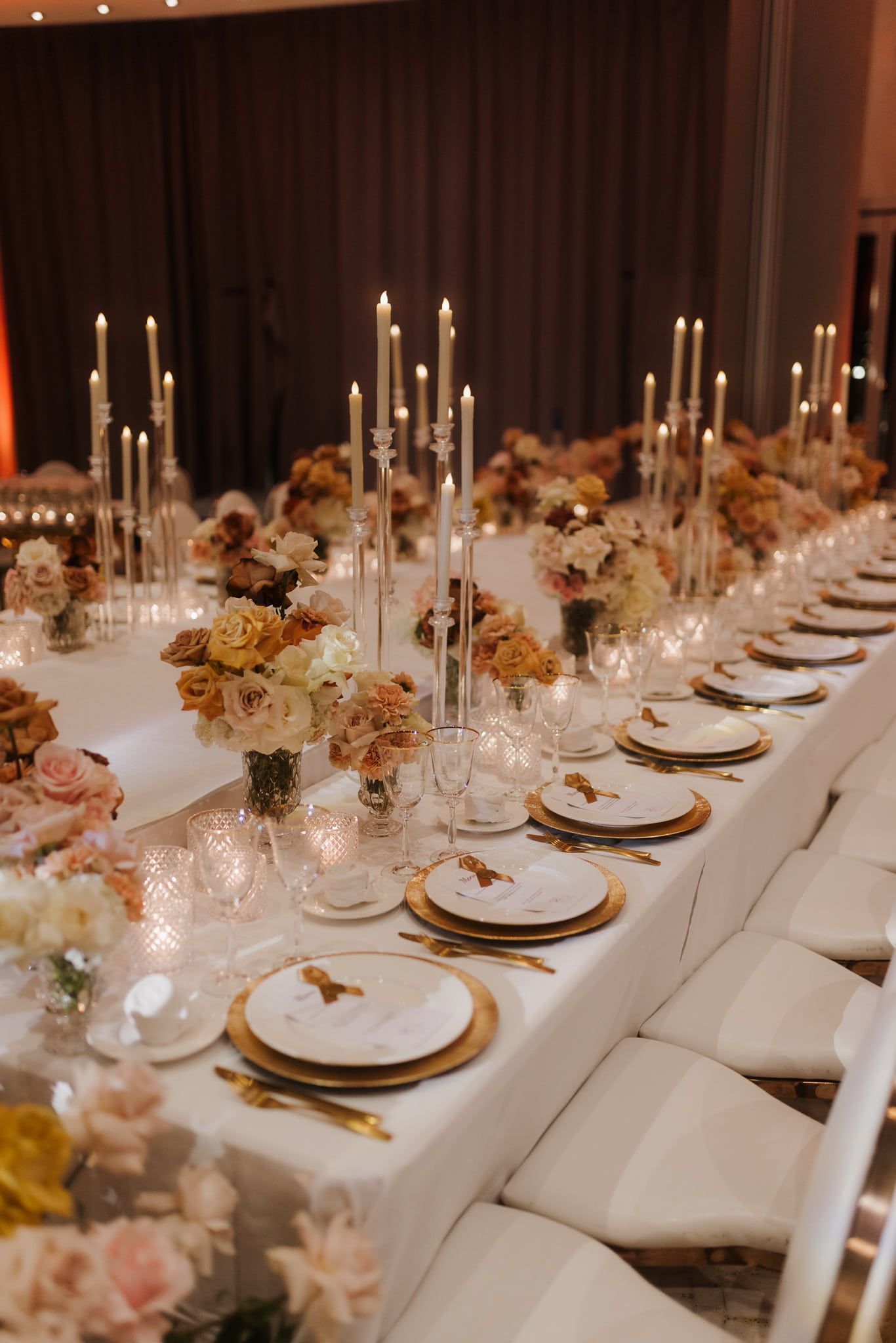 Elegant wedding reception table setting with white linens, gold chargers, and soft pink floral centerpieces lit by glowing candles.