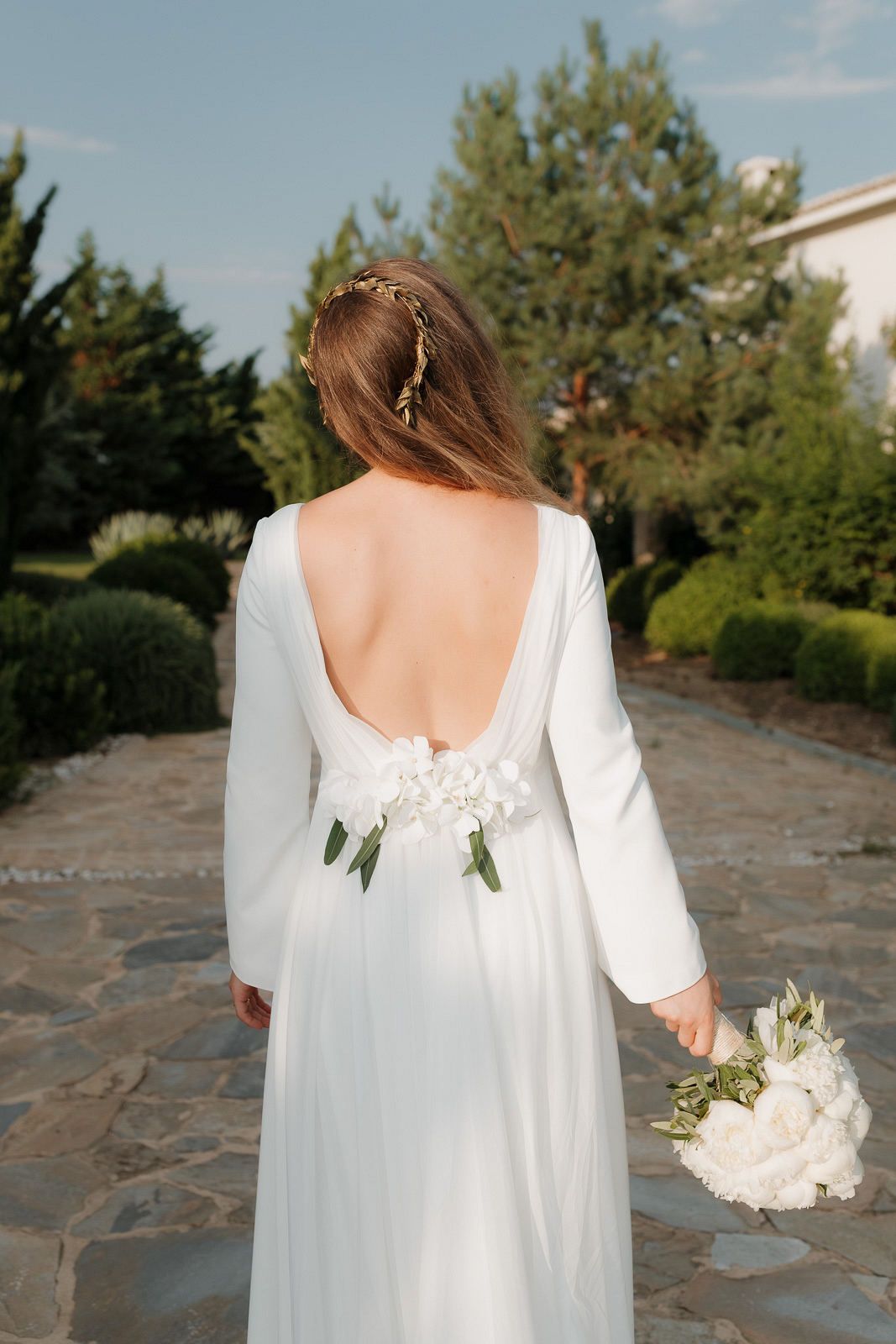 A bride in a flowing white dress walks along a stone path holding a white bouquet, shown from behind.