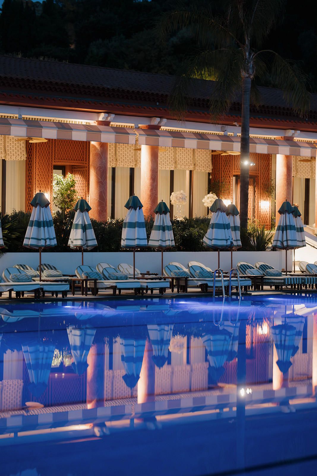 Luxurious resort swimming pool illuminated at night with blue lighting and comfortable lounge chairs along the deck.