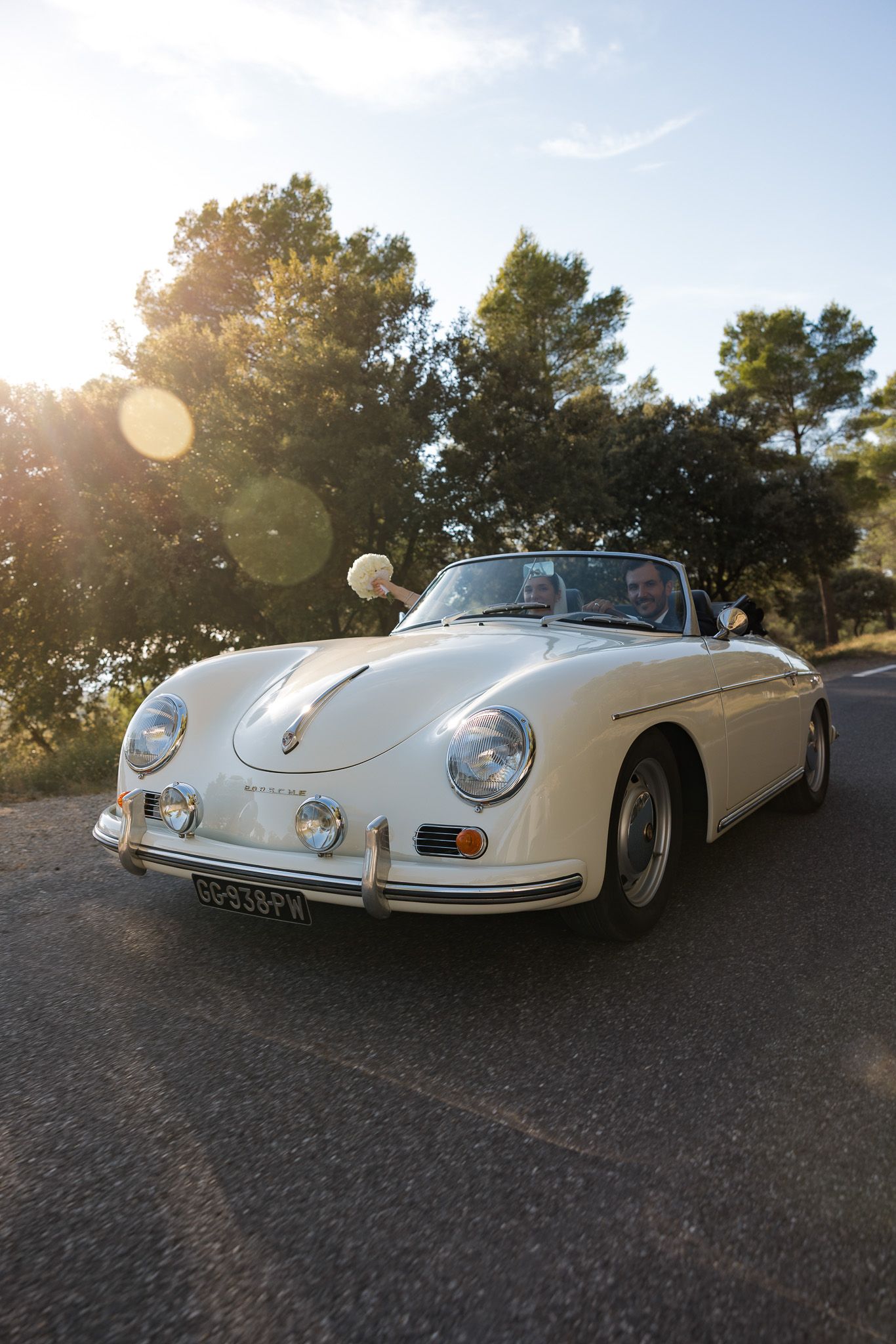 Classic white Porsche 356 Speedster convertible parked on a sunlit road surrounded by lush green trees.
