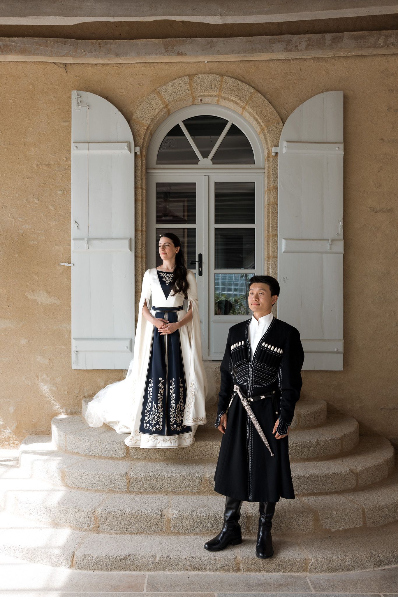 Couple in traditional Georgian wedding attire pose on stone steps before arched white shuttered door.