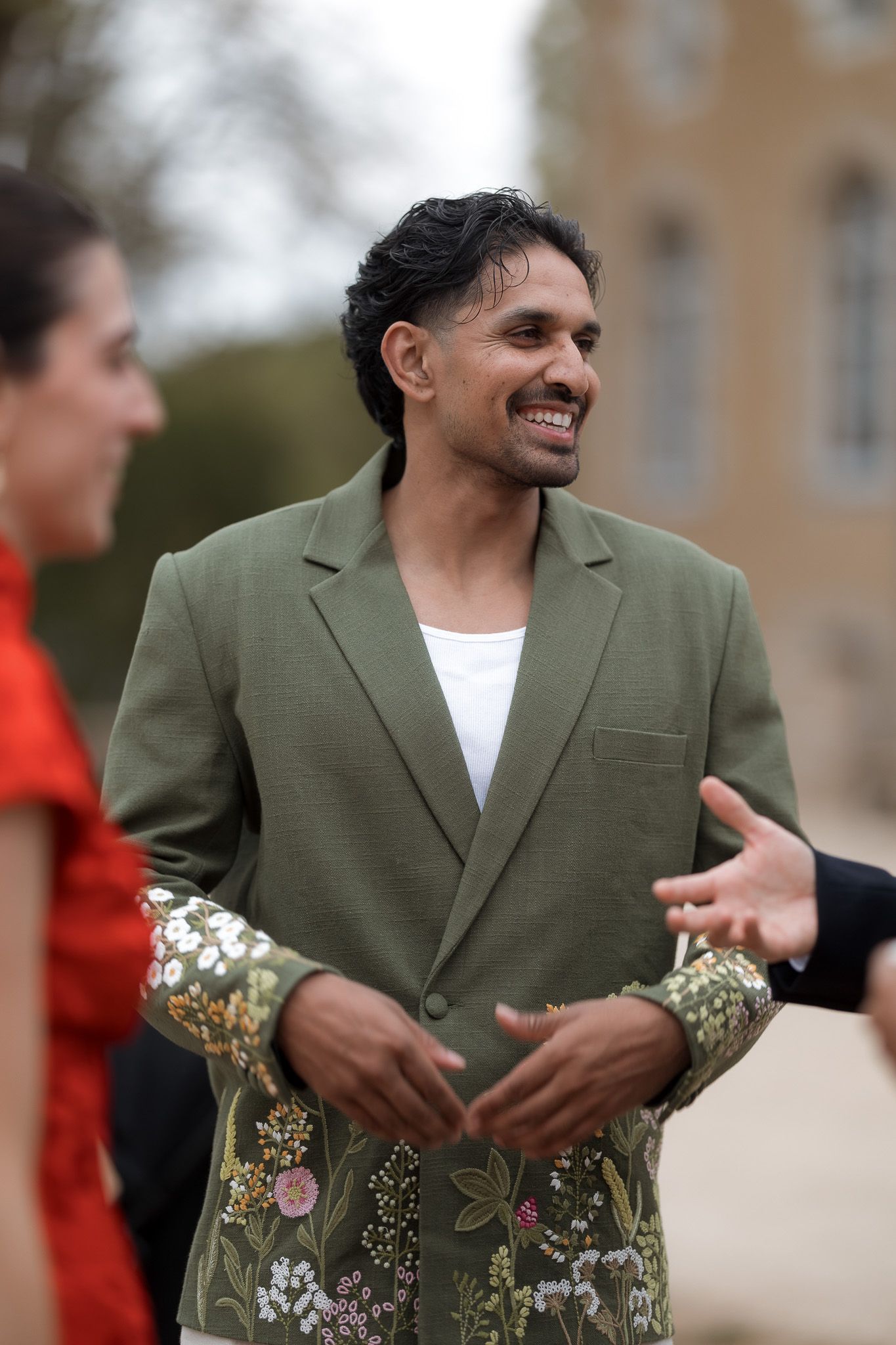 A smiling man in a green blazer with floral embroidered sleeves over a white shirt, socializing outdoors.