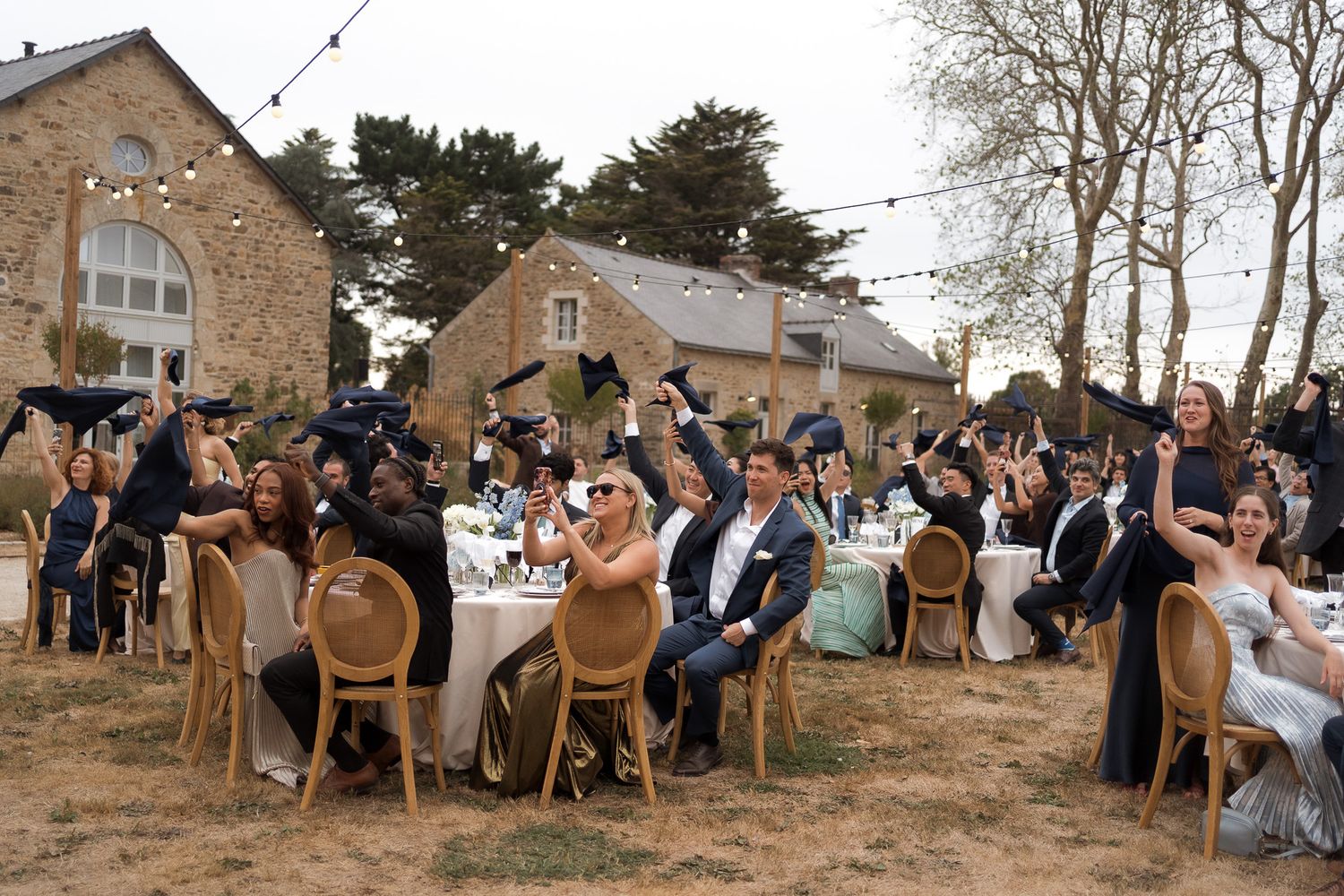 Outdoor wedding reception guests raising glasses in toast at decorated tables near stone building.