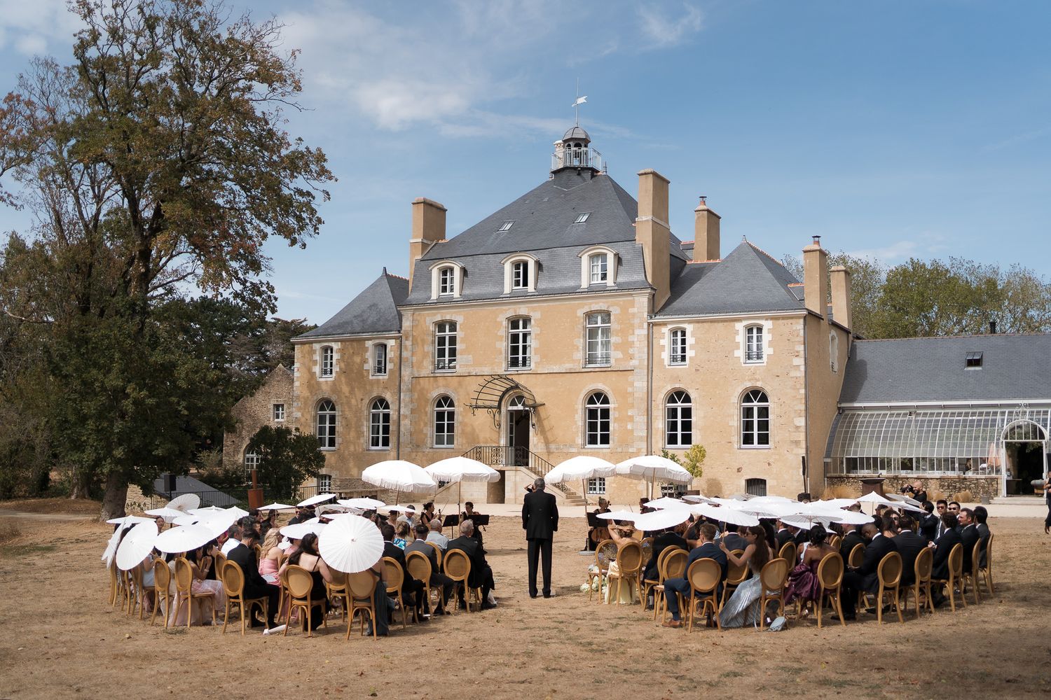 Elegant outdoor wedding ceremony at a French chateau with guests seated under white parasols on a sunny day.