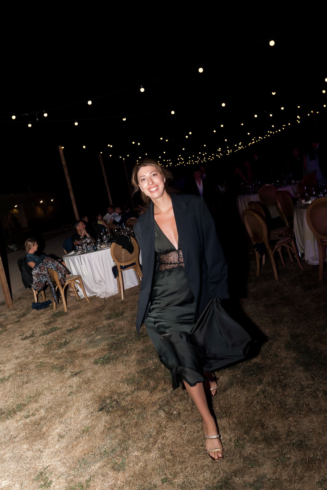 Woman in black blazer and satin dress dancing at an outdoor evening event with string lights overhead.