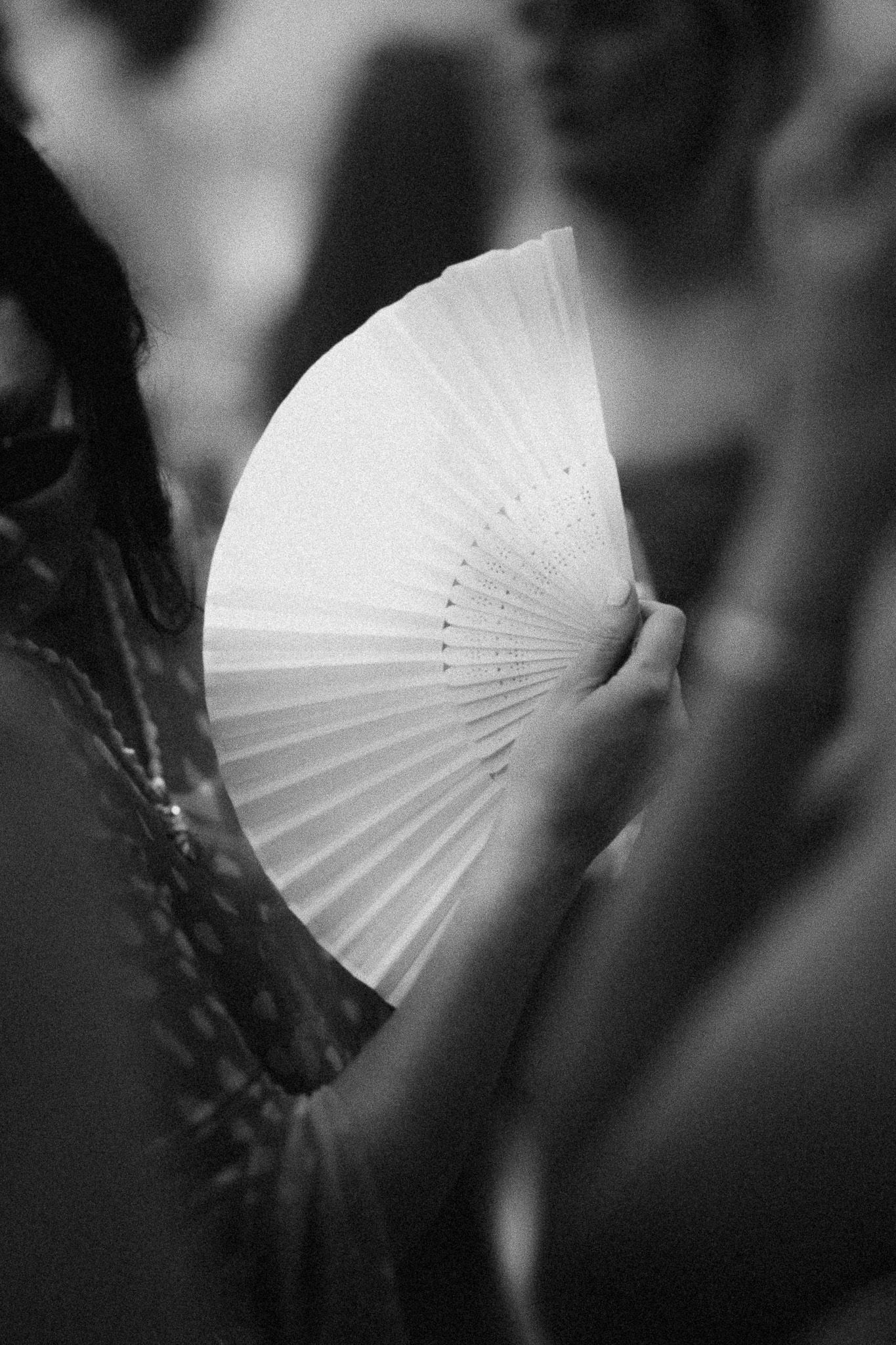 Black and white photo of a woman holding a white folding hand fan at an outdoor event.