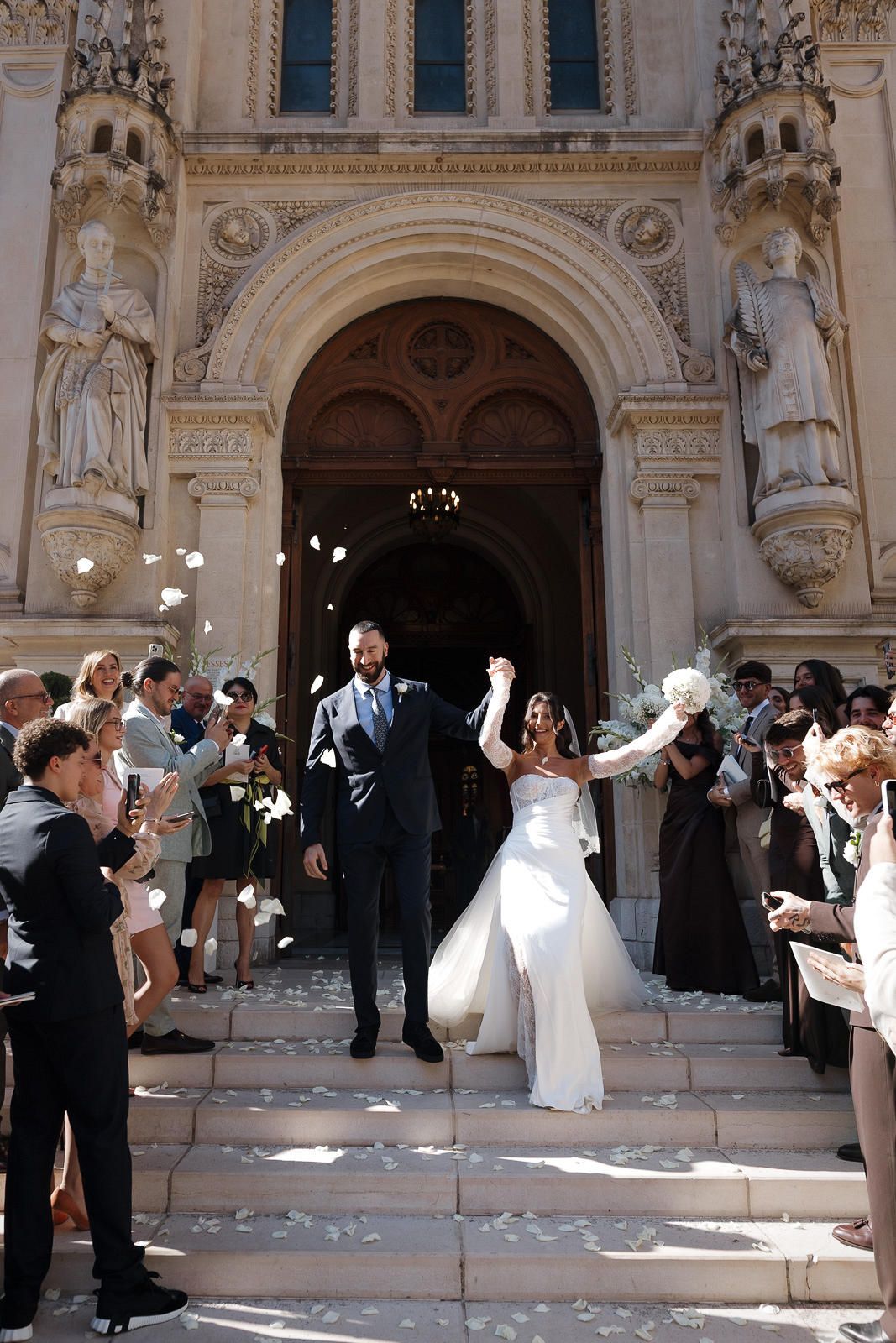 Wedding party celebrates with guests throwing confetti on the steps of an ornate historic building.