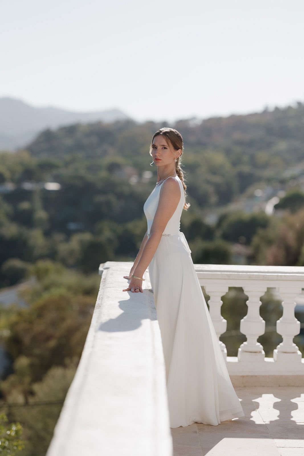 A person in a white wedding dress stands on an elegant balcony overlooking a scenic hillside landscape.