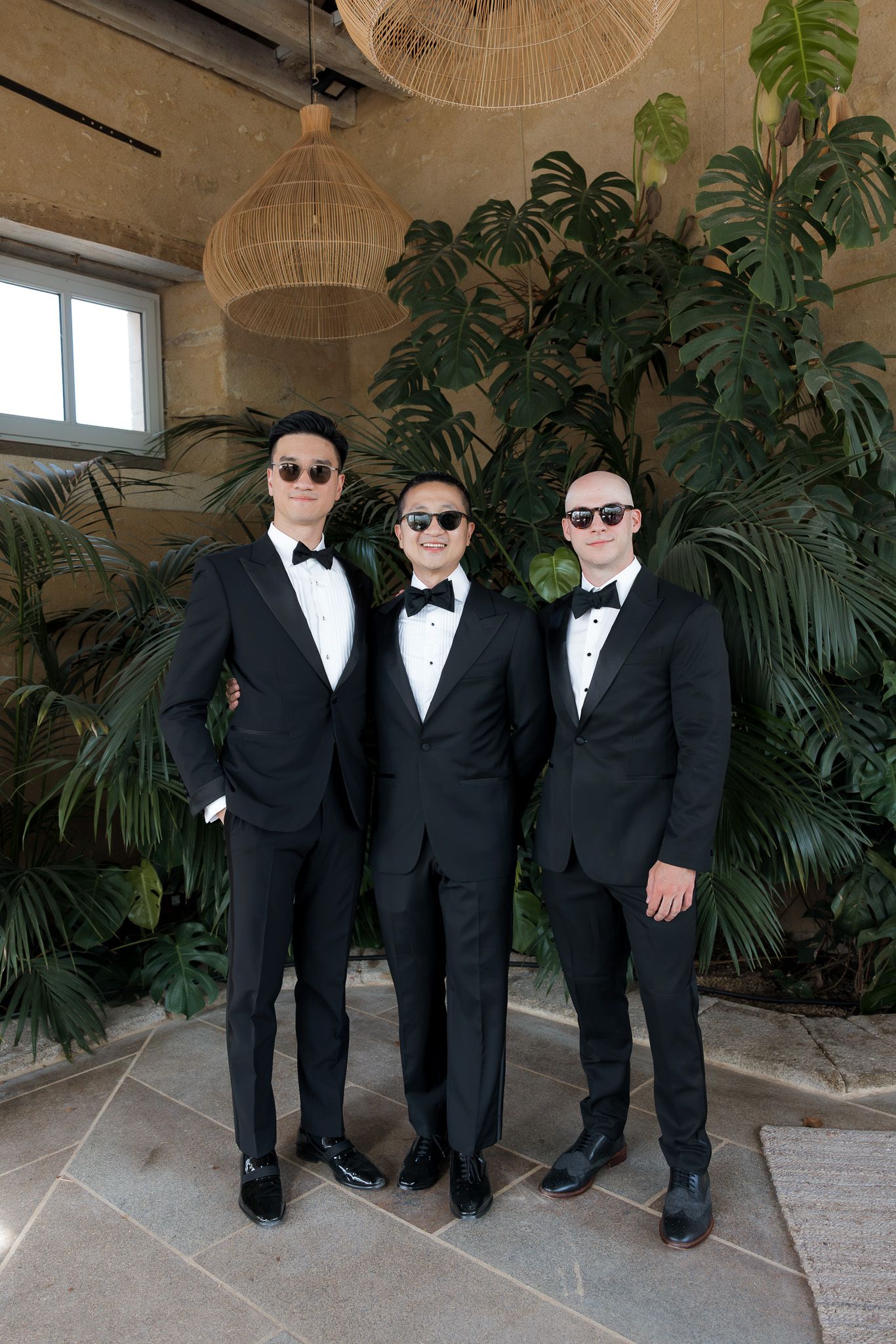 Three groomsmen in black tuxedos and sunglasses pose together in front of tropical greenery at a wedding venue.