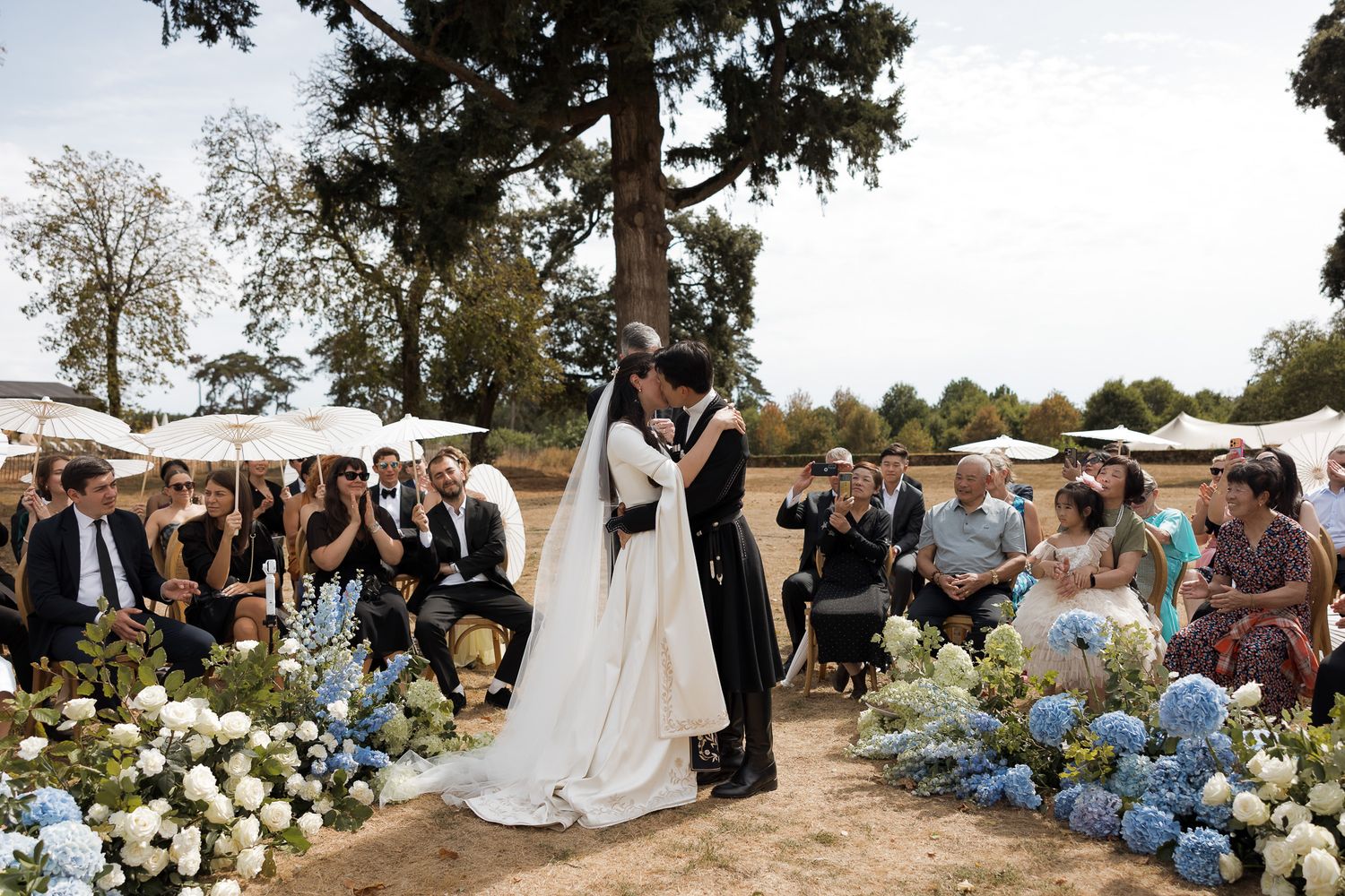 Bride and groom share first kiss at outdoor wedding ceremony lined with blue hydrangeas while guests celebrate.