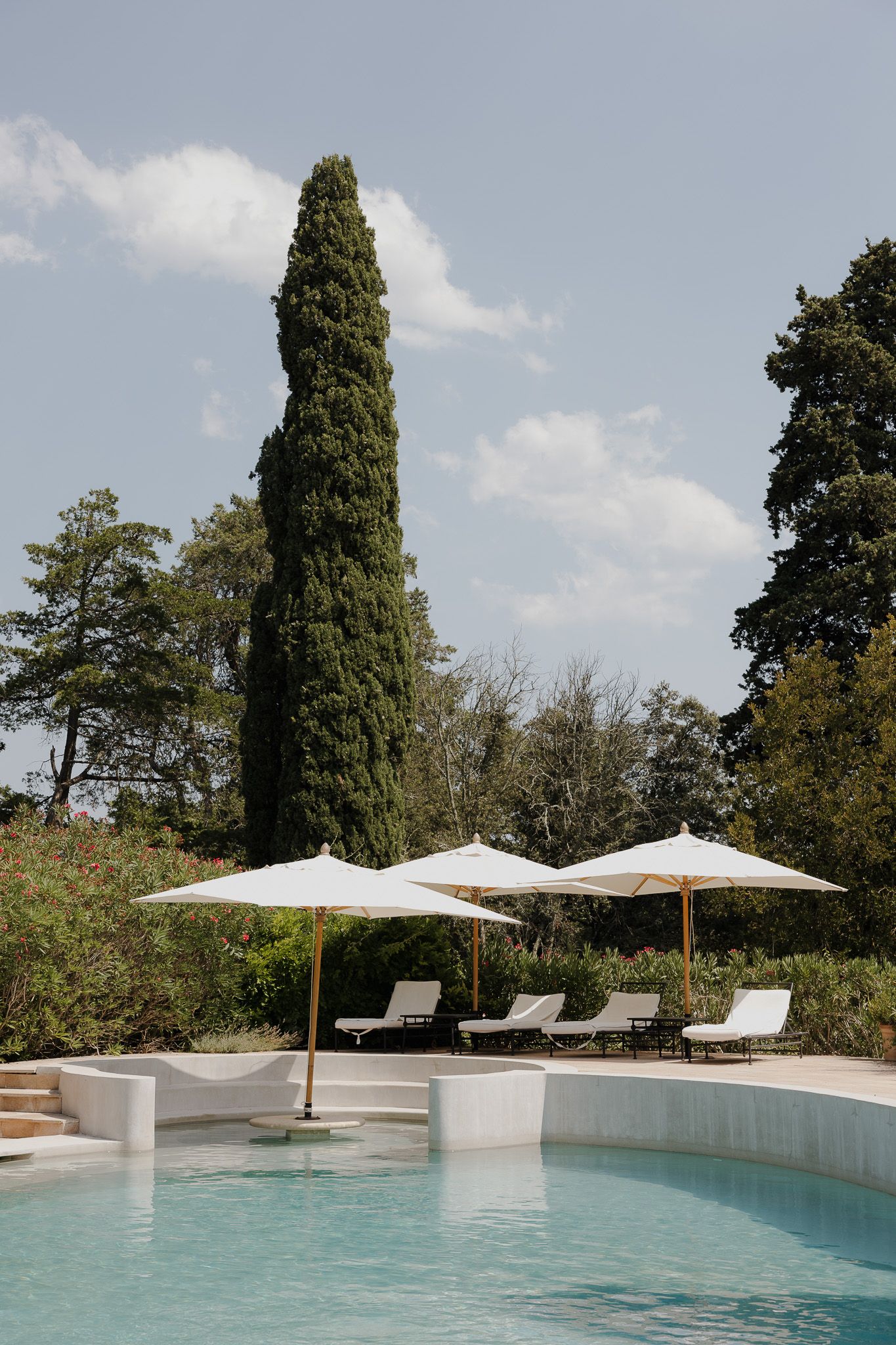 Elegant poolside with white umbrellas, lounge chairs, and a tall cypress tree surrounded by lush greenery under a blue sky.