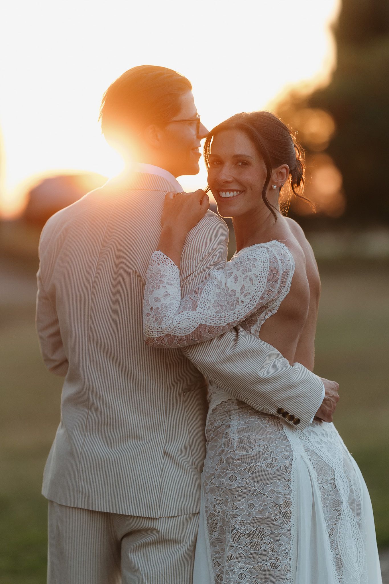 Bride in lace gown embraces groom in tan suit during golden sunset, smiling warmly at camera.