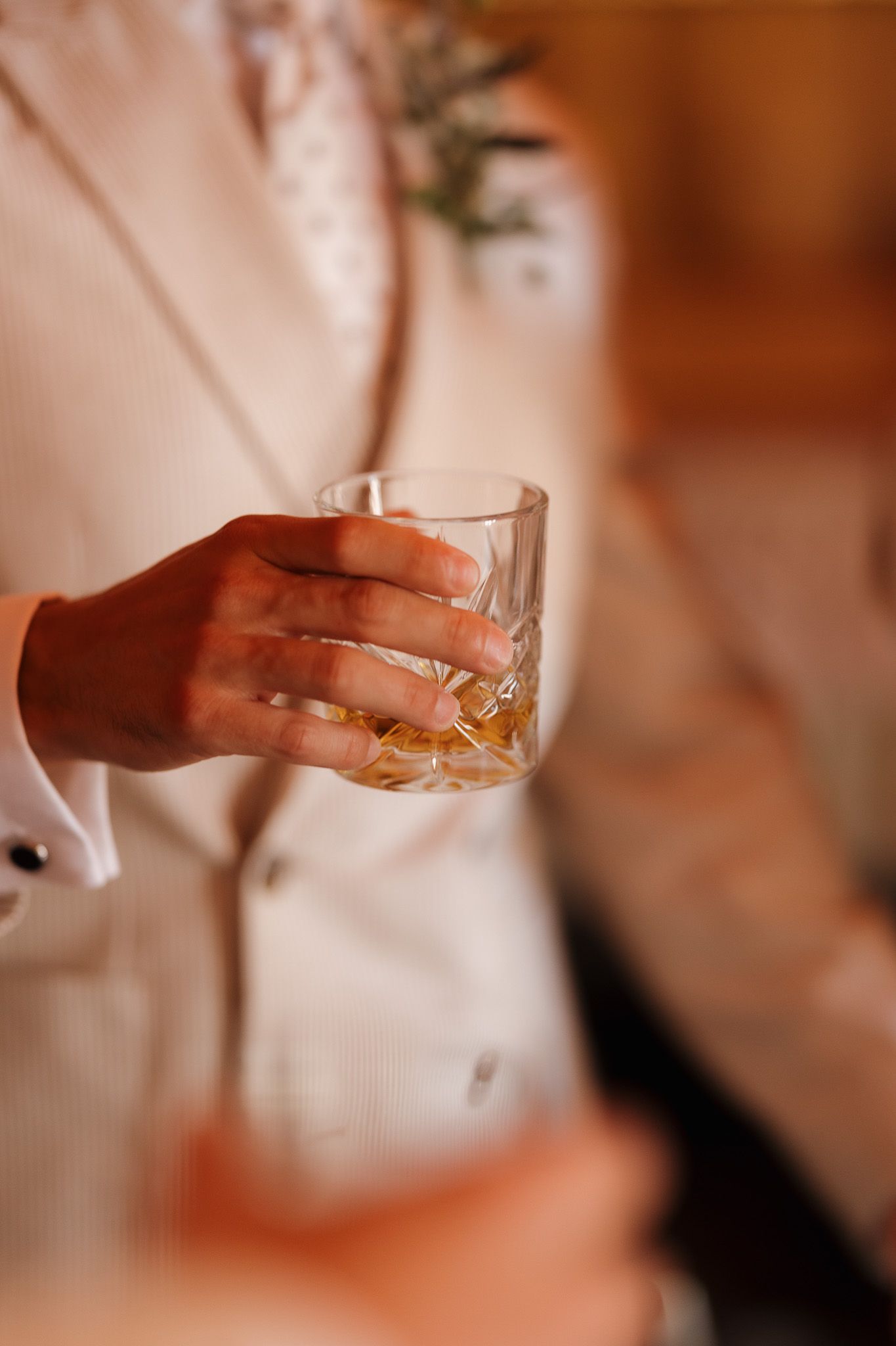 Person in white shirt holding a crystal whiskey glass with amber liquid at a formal event.