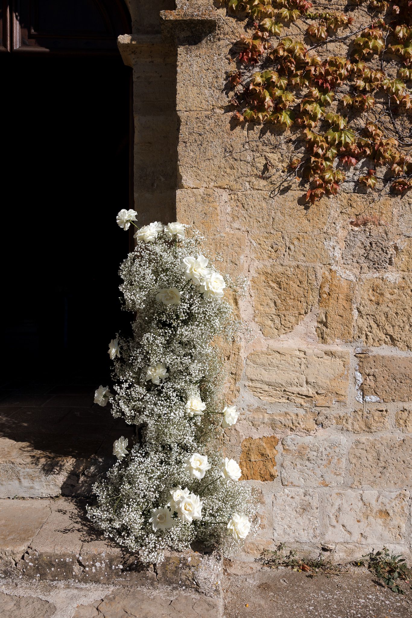 White roses and baby's breath floral arrangement decorating a rustic stone church entrance doorway.