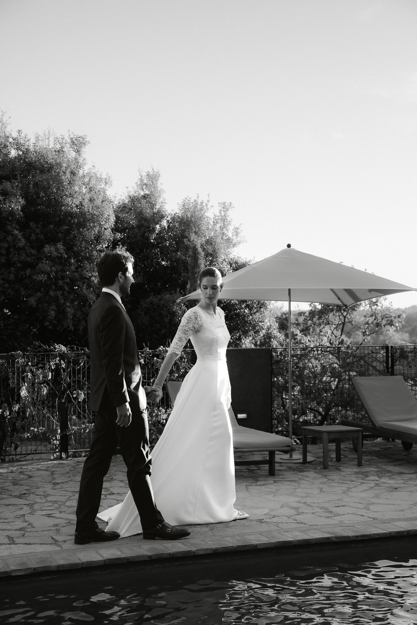 Black and white photo of a bride and groom walking hand in hand beside a pool at an outdoor wedding venue.