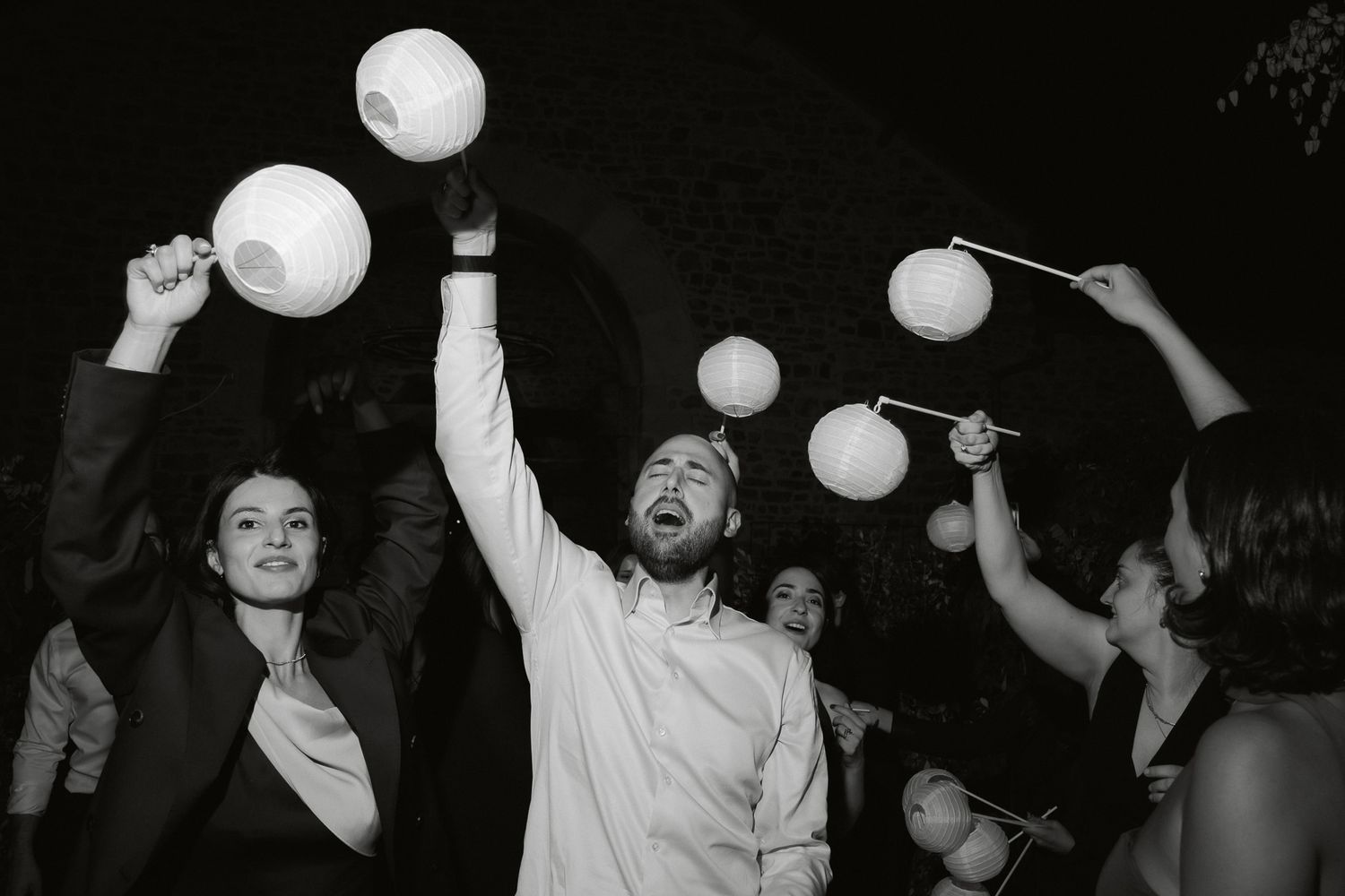 Black and white photo of joyful wedding guests dancing and holding paper lanterns at a lively reception celebration.