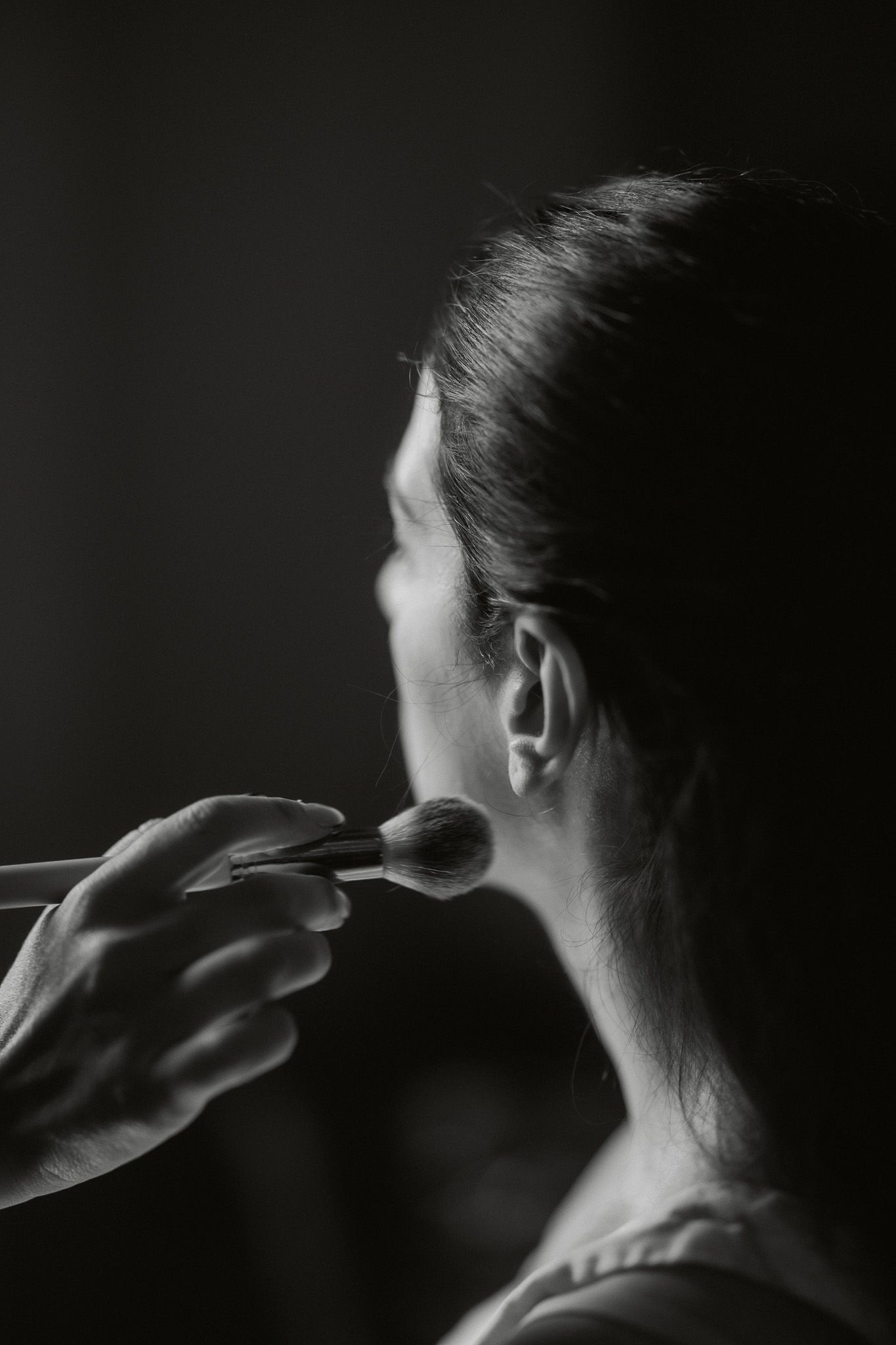 Black and white photo of a makeup artist applying blush to a womans cheek with a brush in a studio.