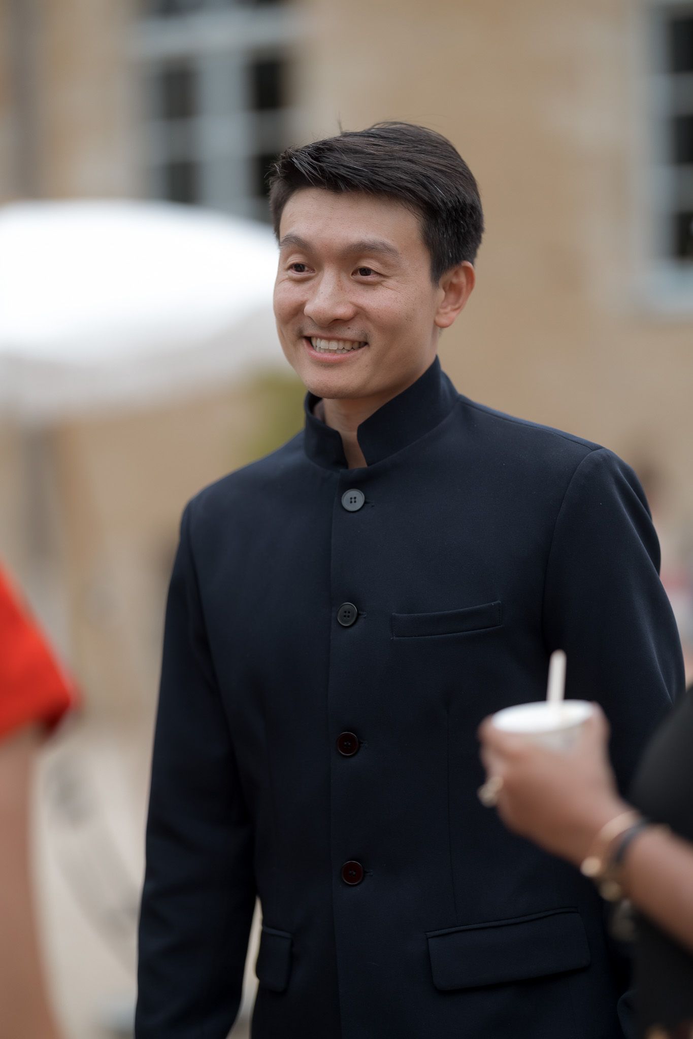 Smiling Asian man in a dark navy Mandarin collar jacket holding a cup at an outdoor event.