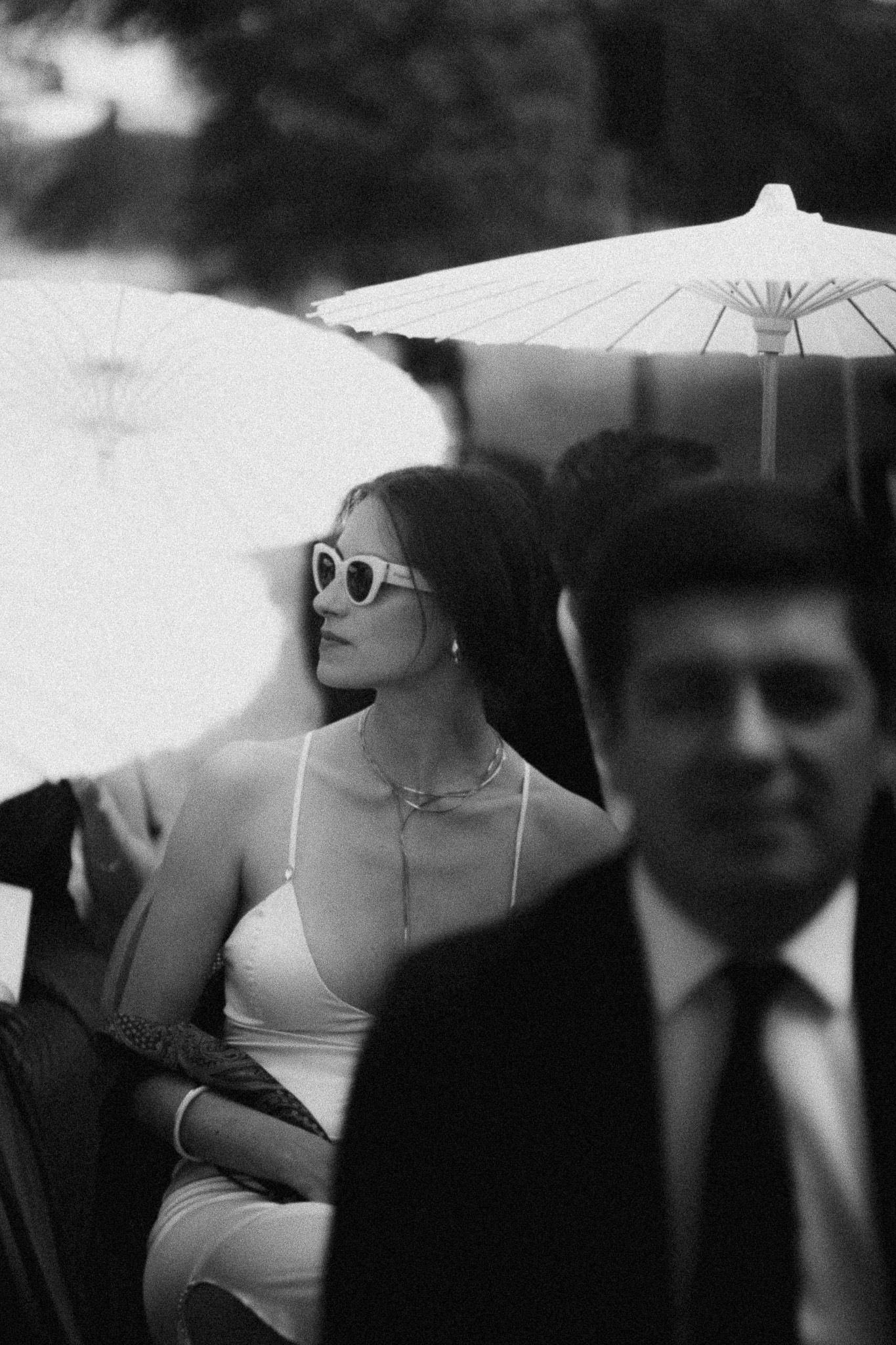 Black and white photo of elegant woman in white dress and sunglasses under parasol at formal outdoor event.