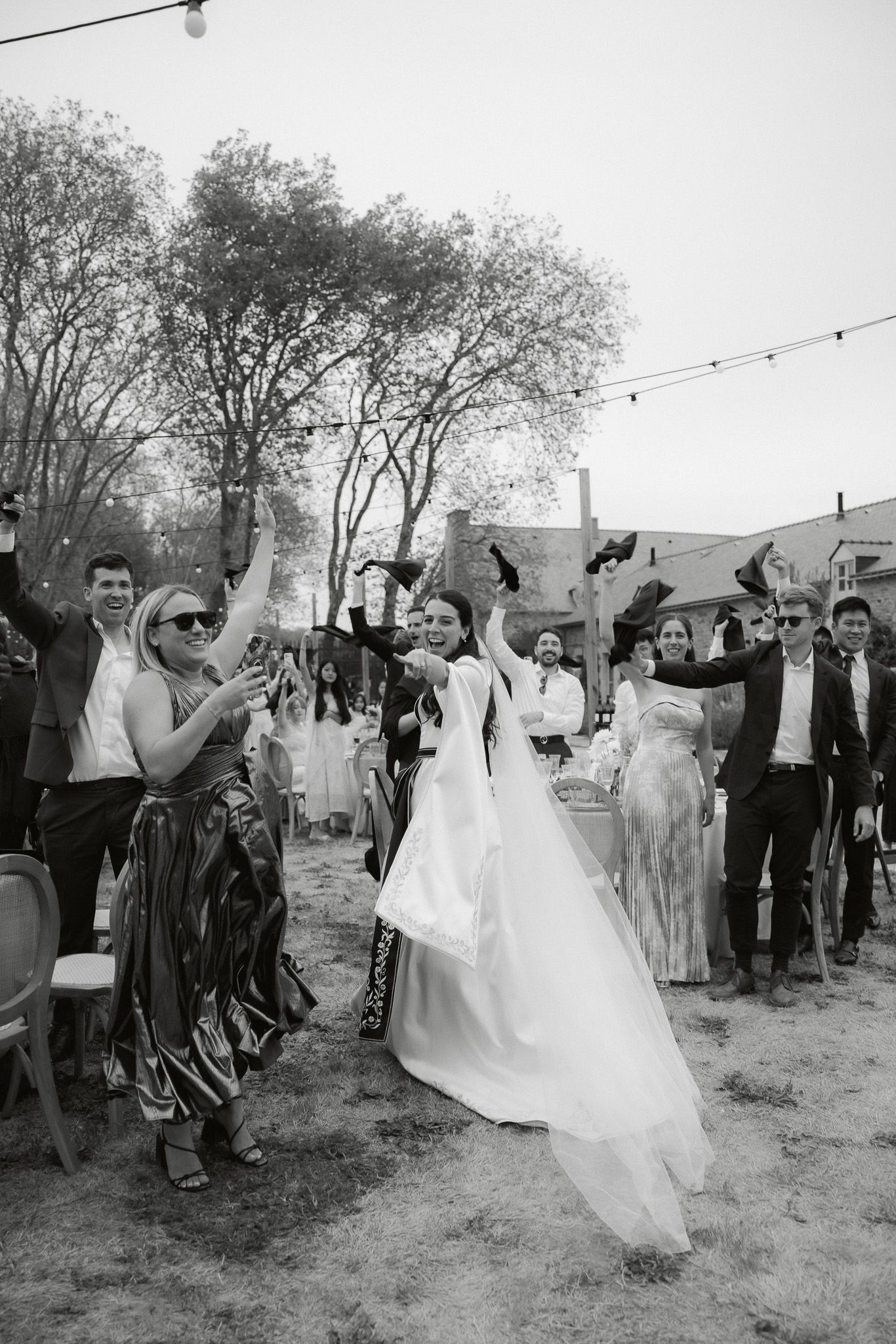Black and white photo of a joyful bride in white gown celebrating with guests outdoors at a wedding reception.