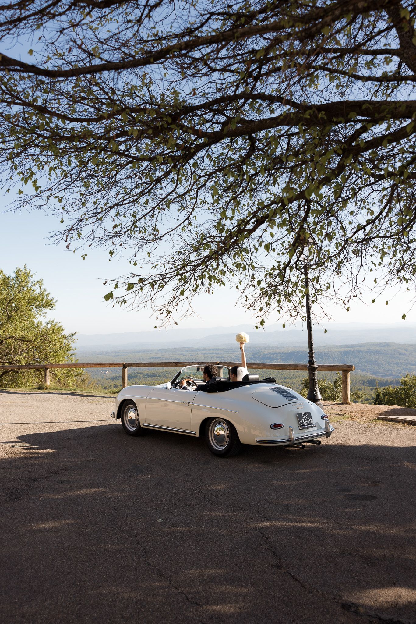 Couple in a vintage white Porsche 356 convertible at a scenic overlook, framed by tree branches.