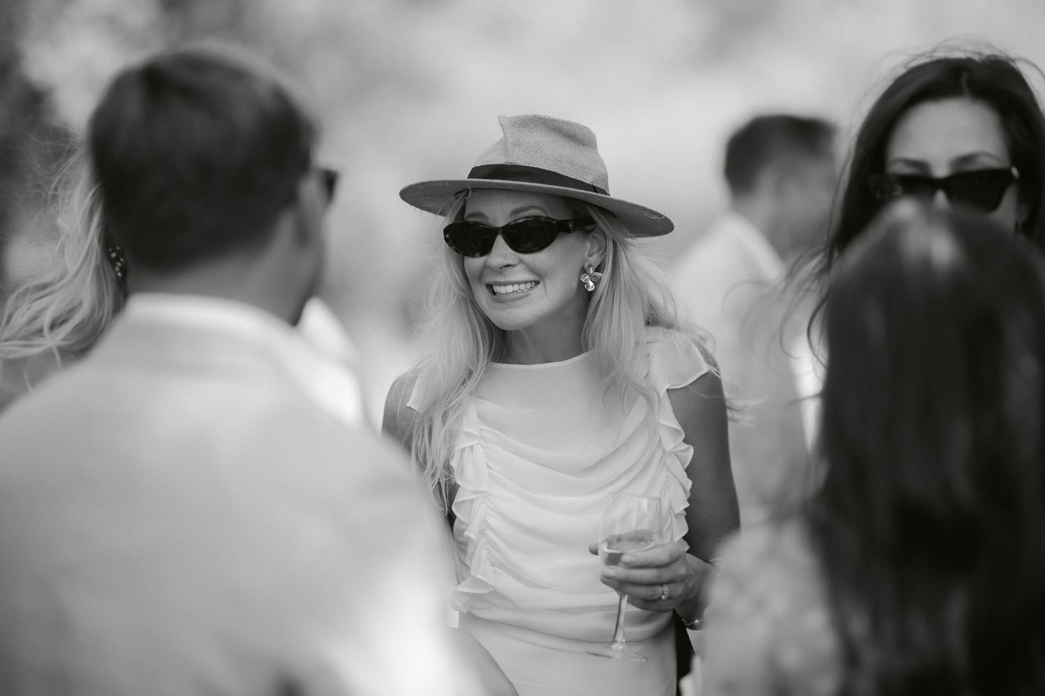 A smiling woman in a wide-brim hat and sunglasses enjoys a social gathering in this elegant black and white photo.
