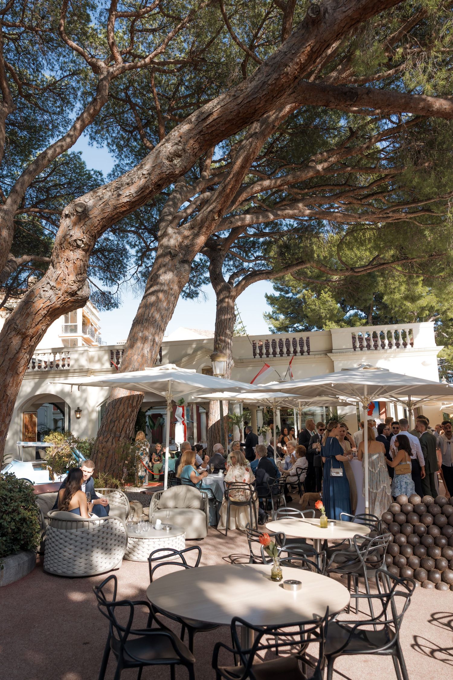 Elegant outdoor terrace cafe under large pine trees with guests gathered near a white Mediterranean villa.