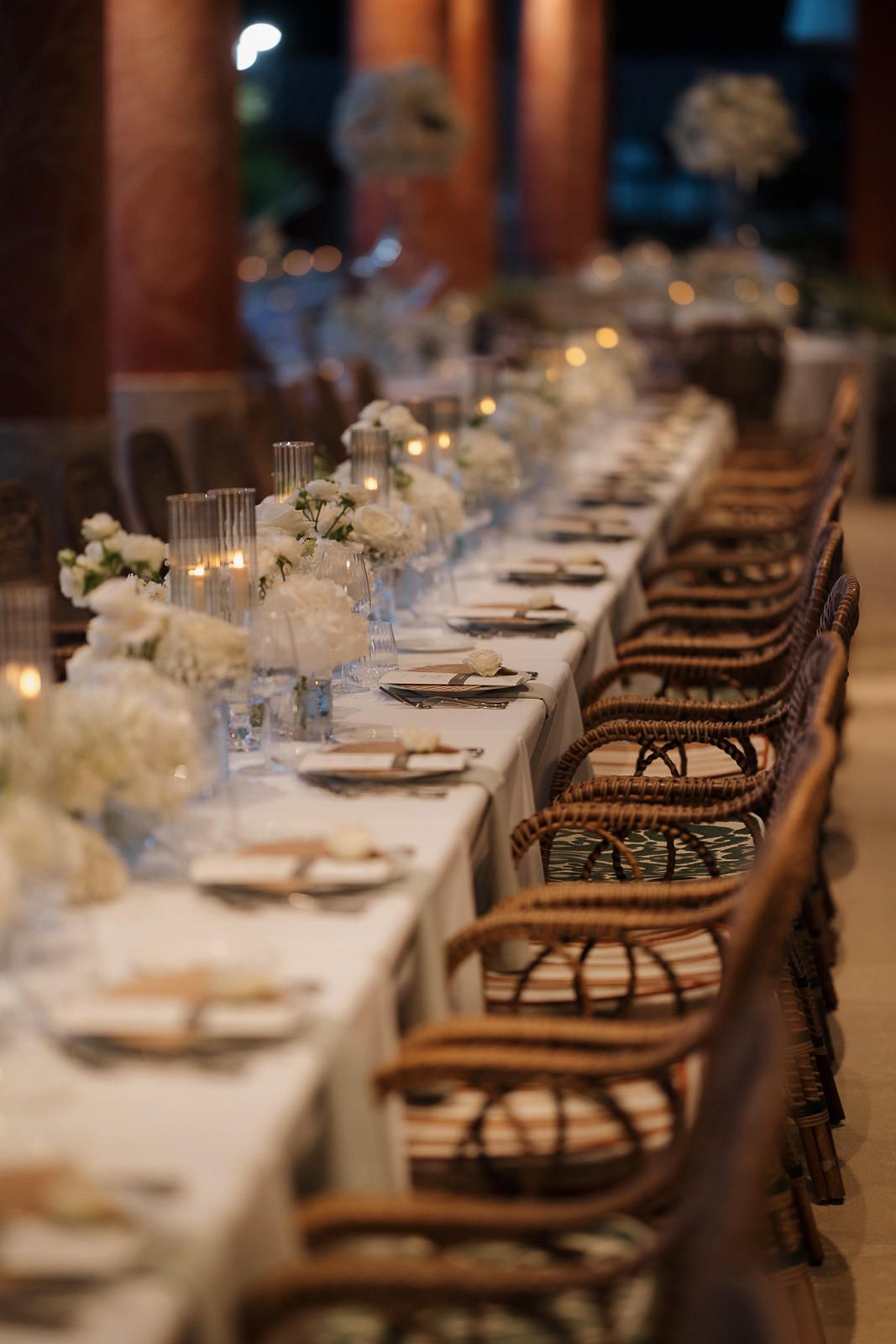 Elegant wedding reception table setting with white linens, ornate chairs, and floral centerpieces in warm ambient lighting.