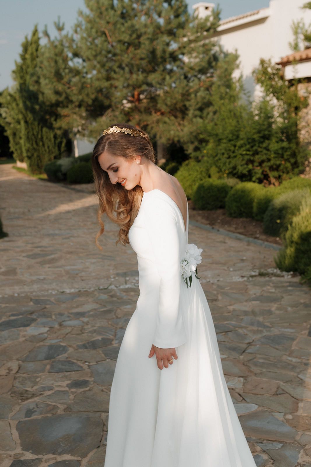 A person in a flowing white wedding dress spins gracefully on a stone patio surrounded by greenery and buildings.
