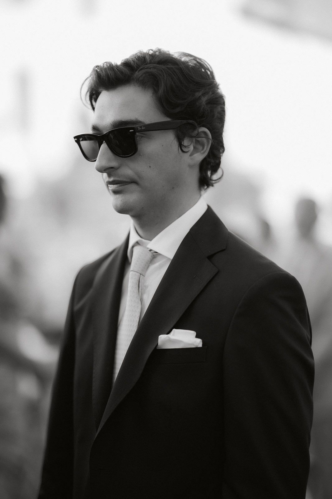 A black and white portrait of someone wearing sunglasses, a suit and tie with a pocket square.