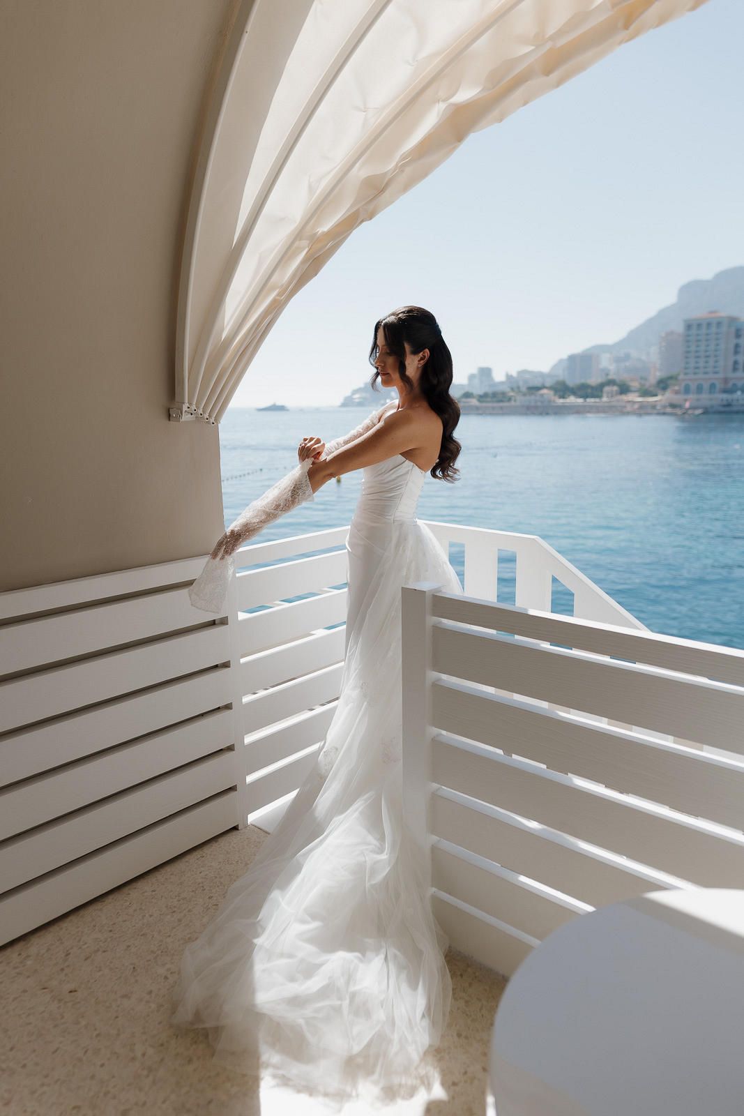 Elegant white wedding dress flowing in the breeze on a seaside balcony overlooking blue Mediterranean waters.