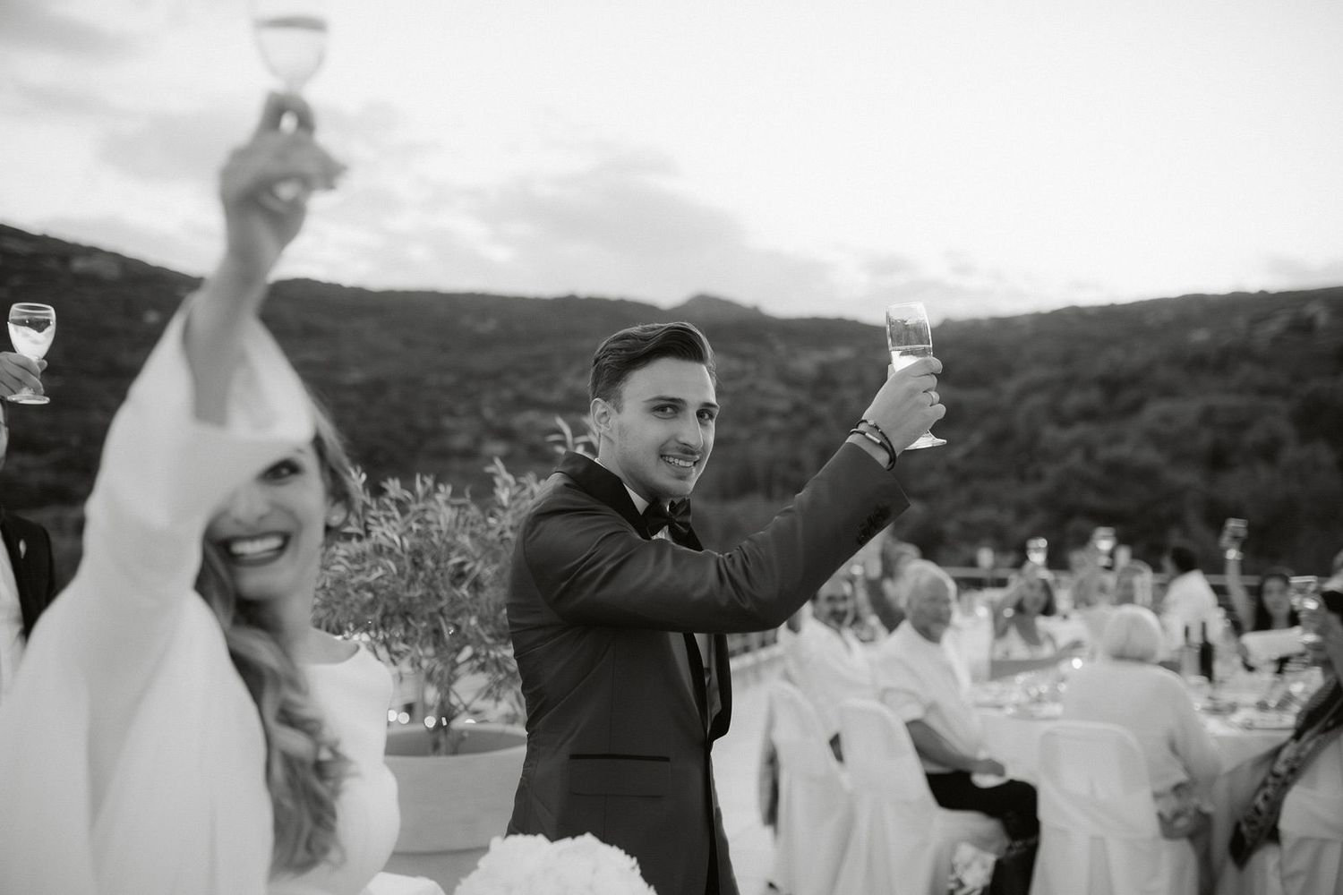 Wedding guests celebrate with raised arms during an outdoor reception in a scenic mountain setting.