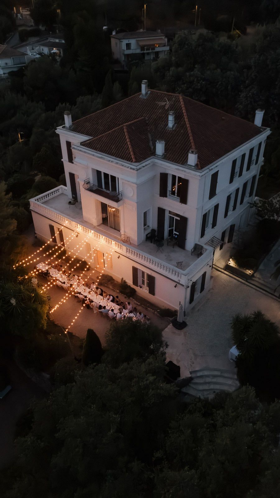 Aerial view of an elegant white mansion with outdoor dining tables illuminated by string lights at dusk.