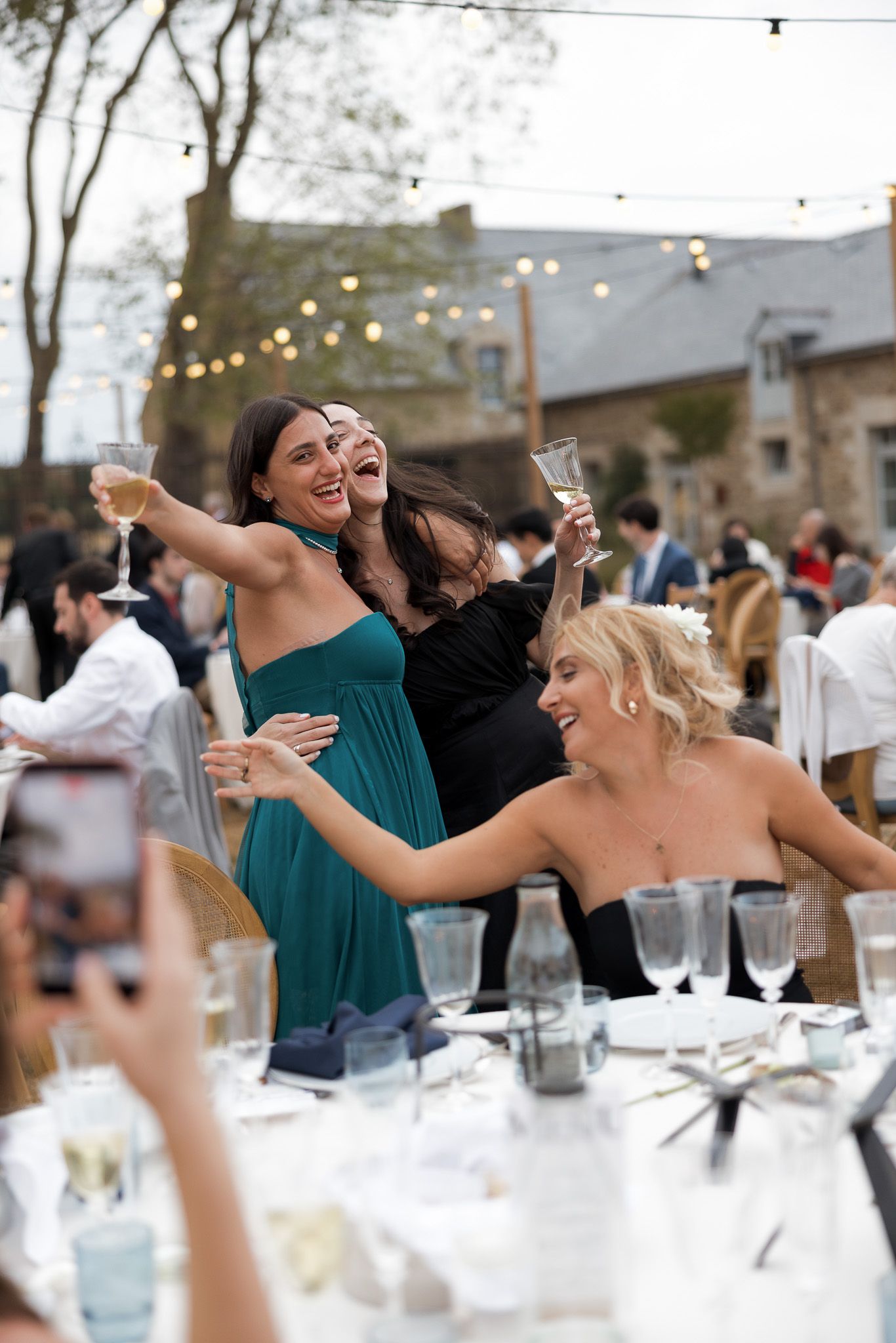 Three women laughing and raising champagne glasses at an outdoor wedding reception dinner.