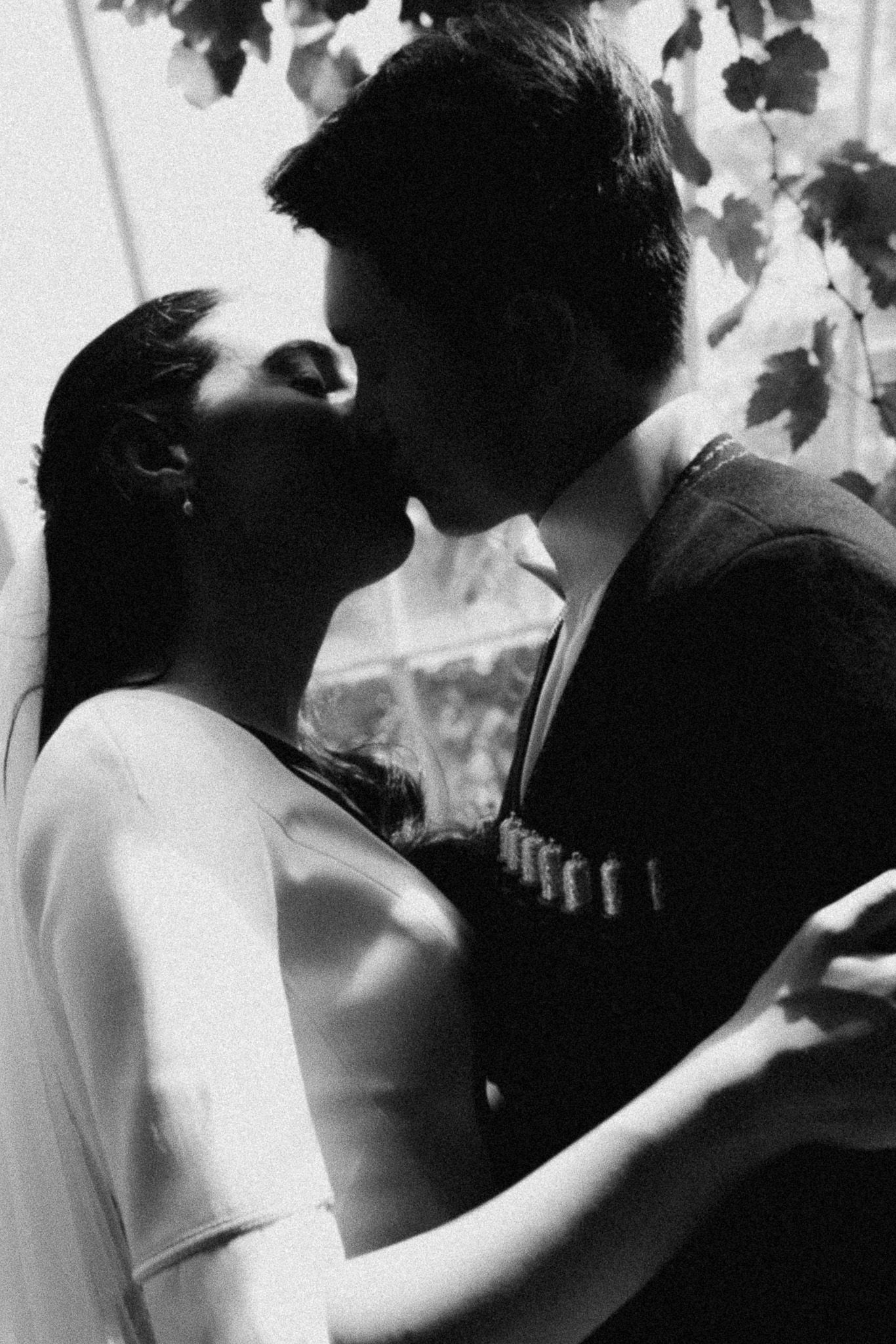 Black and white photo of a couple sharing a romantic kiss outdoors beneath leafy branches.