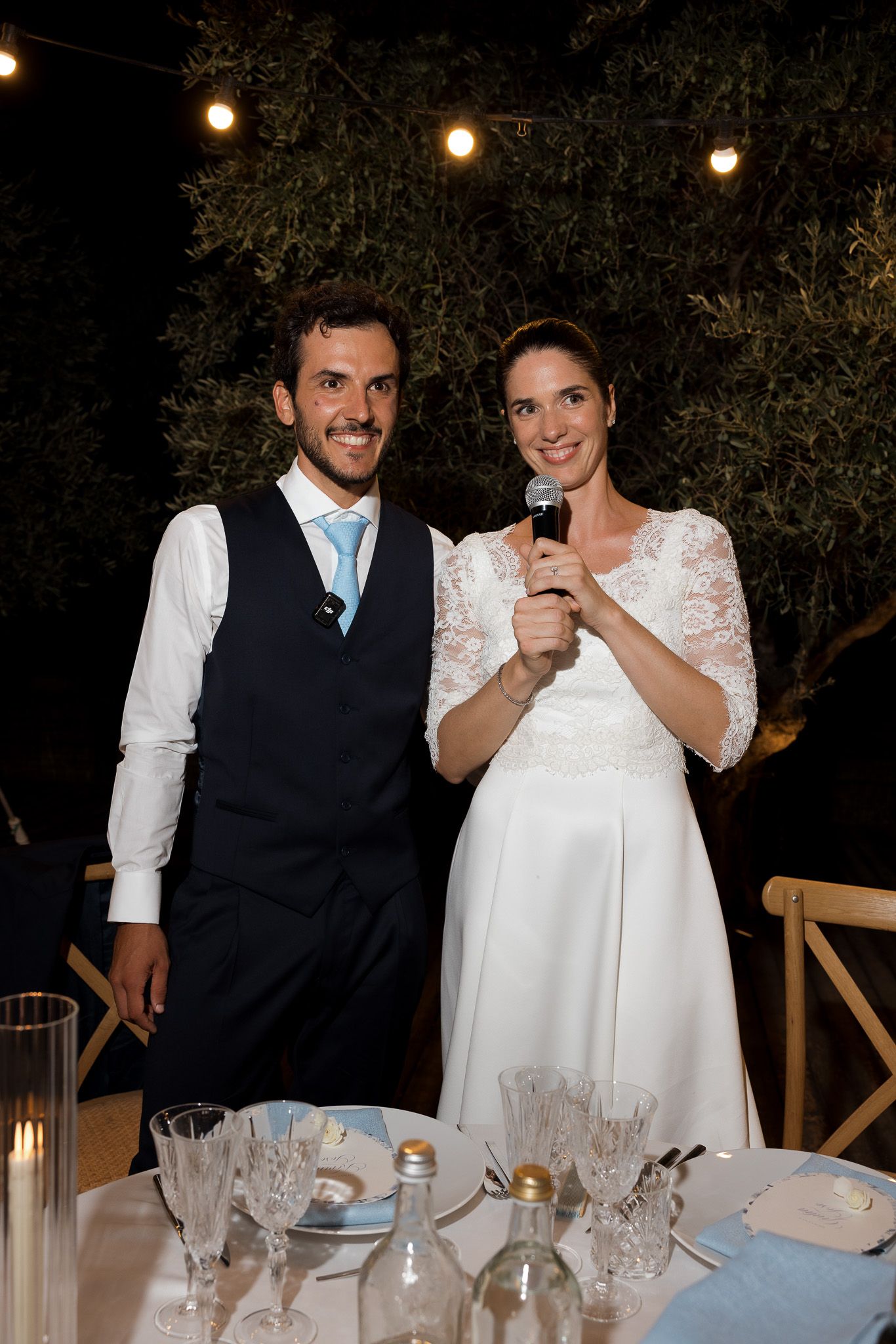 A smiling bride and groom pose together at their outdoor wedding reception, bride holding a microphone.