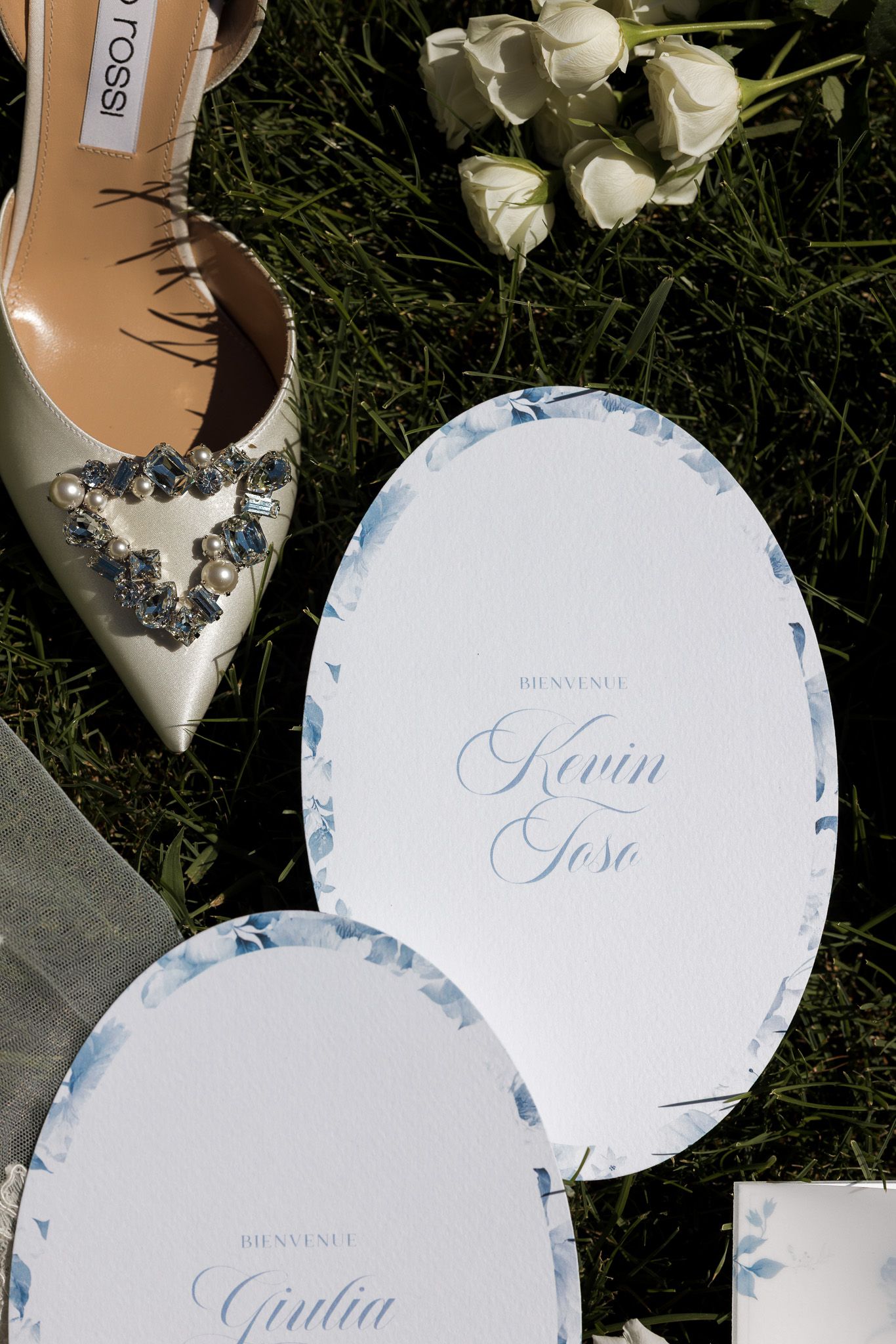 Elegant wedding stationery with blue floral details, personalized name cards, and a jeweled bridal heel on green grass.