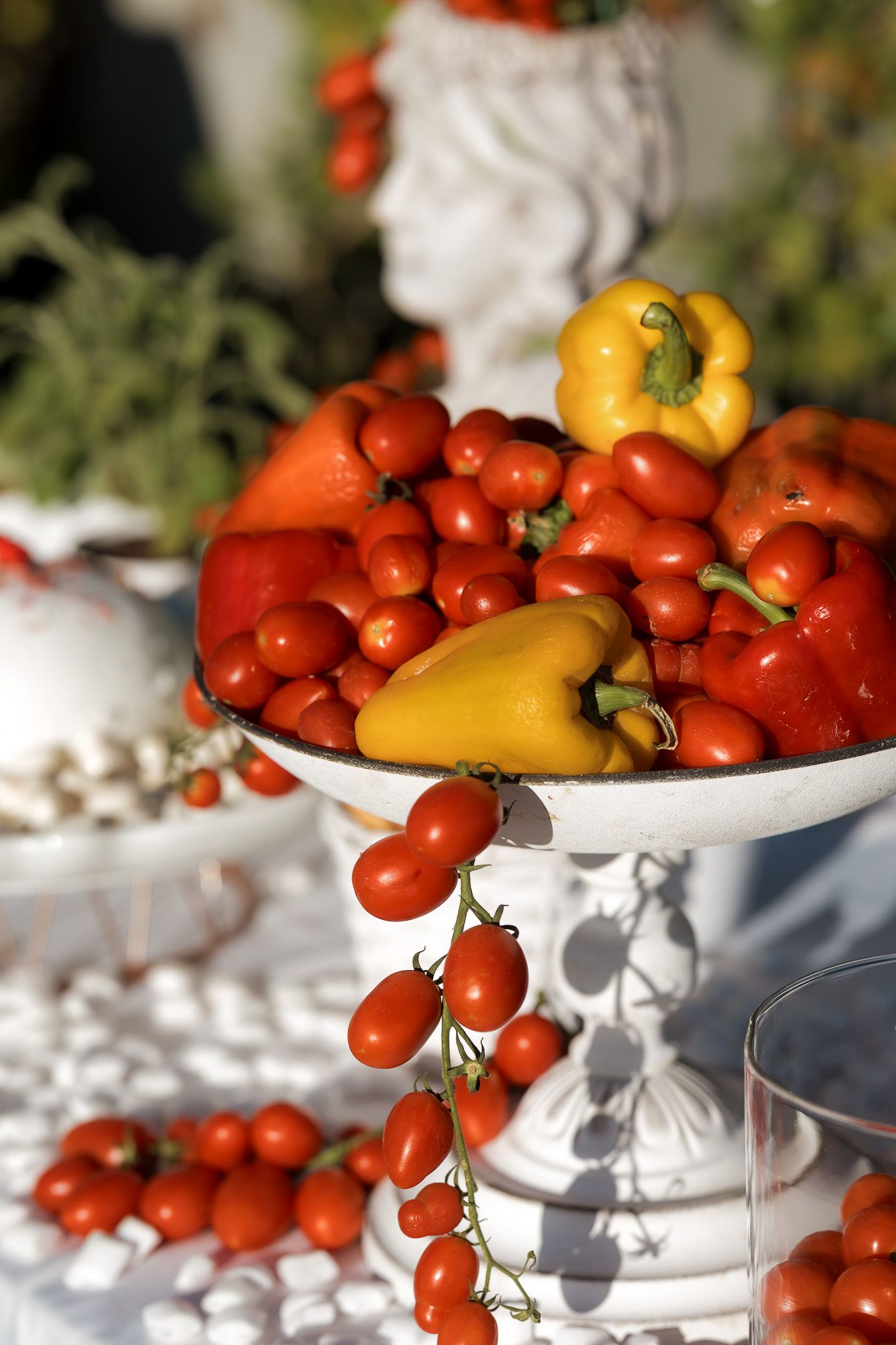 White pedestal bowl filled with cherry tomatoes and yellow bell peppers displayed outdoors on a white tablecloth.