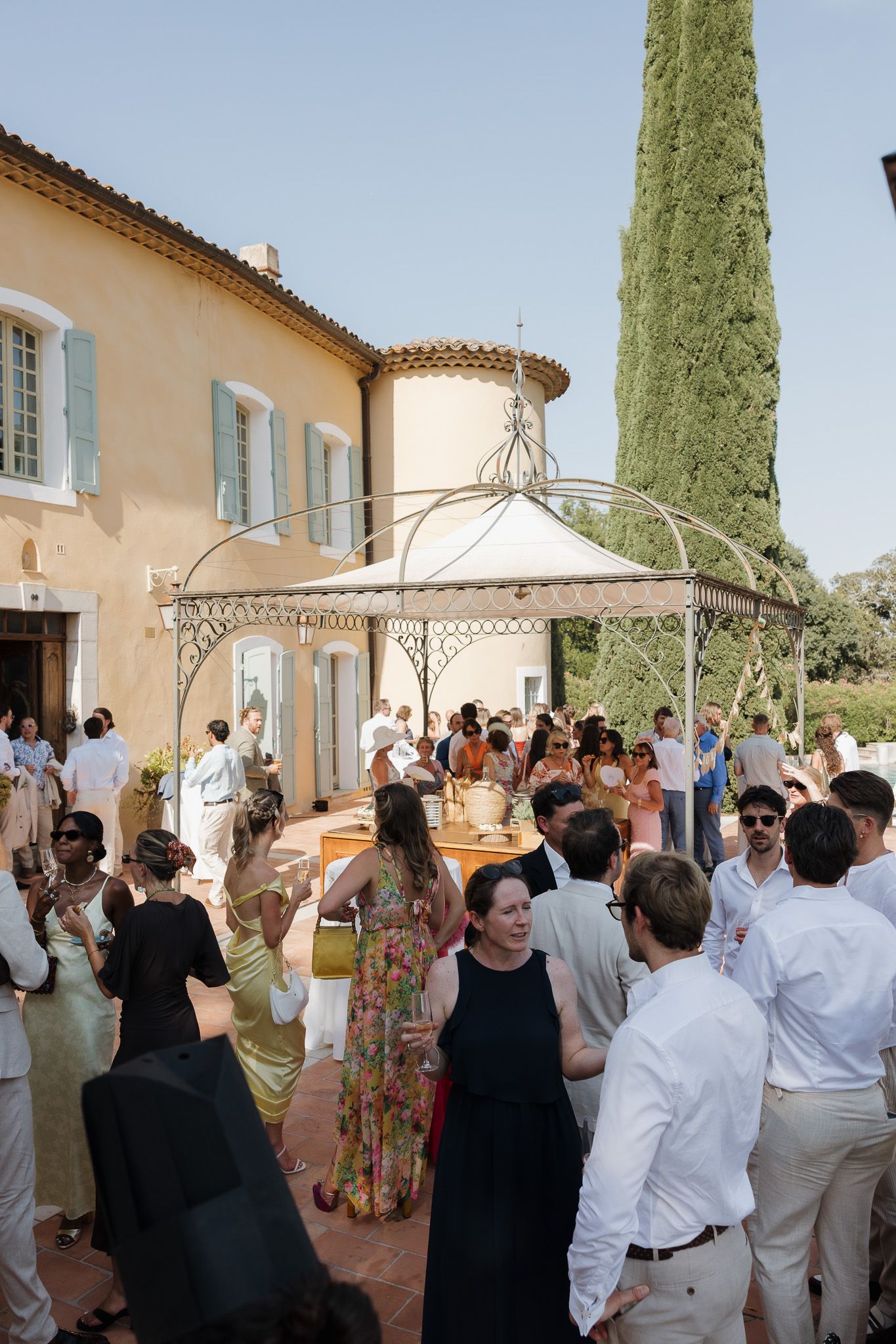 Elegant outdoor wedding reception at a Provençal villa with guests mingling under an ornate gazebo on a sunny day.