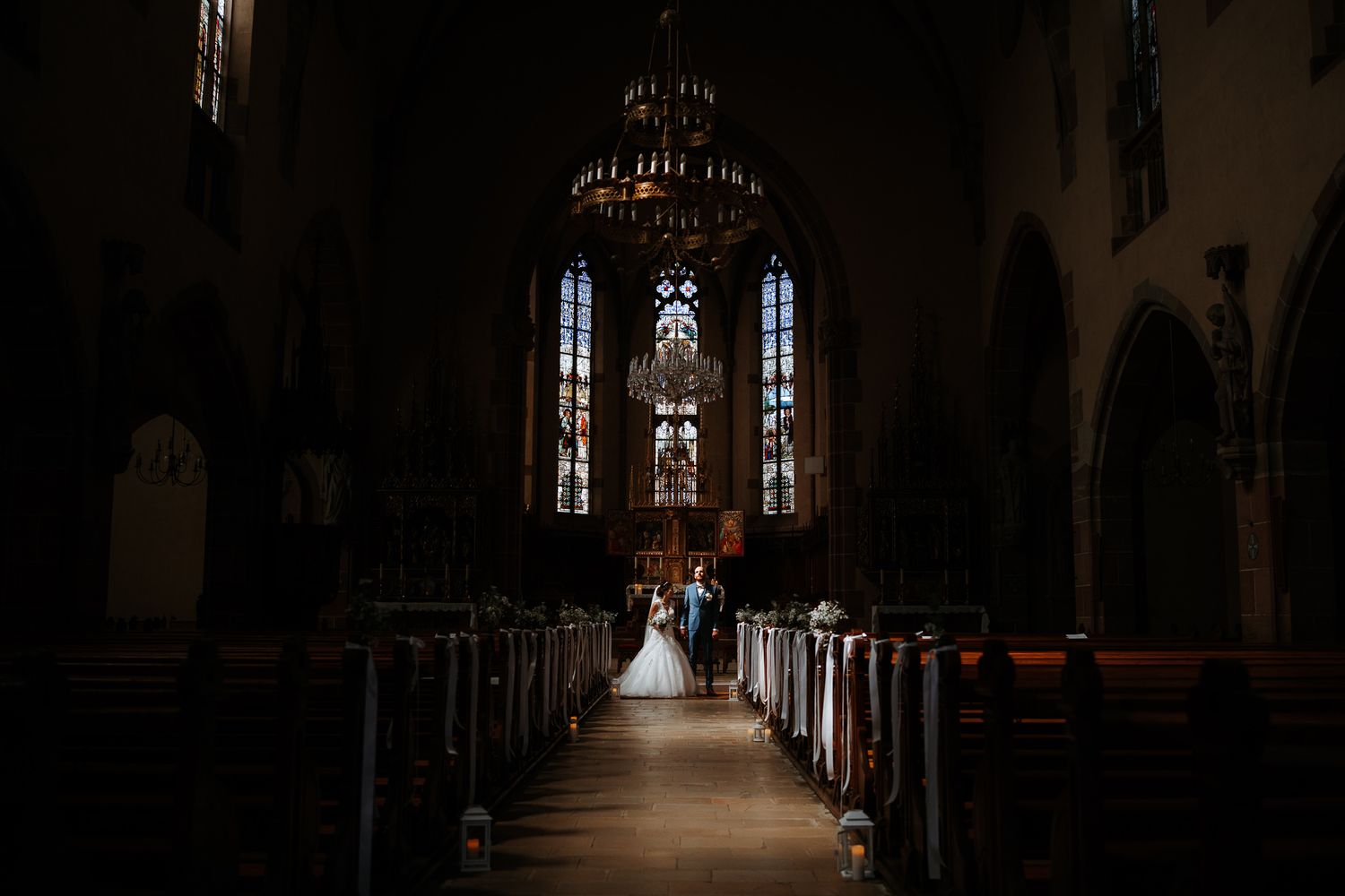 A long church aisle with stained glass windows leads to a couple getting married at the altar in a dimly lit cathedral.