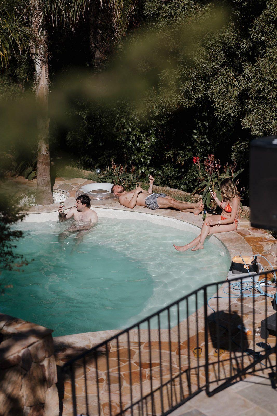 People relaxing by a luxurious swimming pool surrounded by lush greenery and palm trees on a sunny day.
