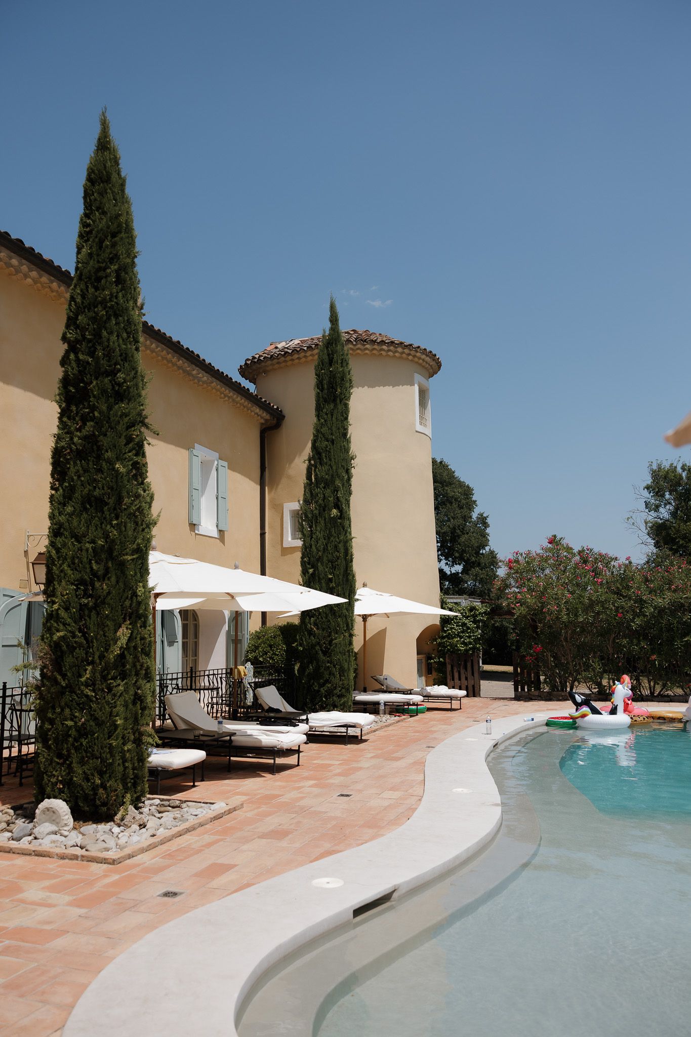 Elegant Provençal villa with cypress trees, lounge chairs, white umbrellas, and a curved swimming pool under clear blue sky.