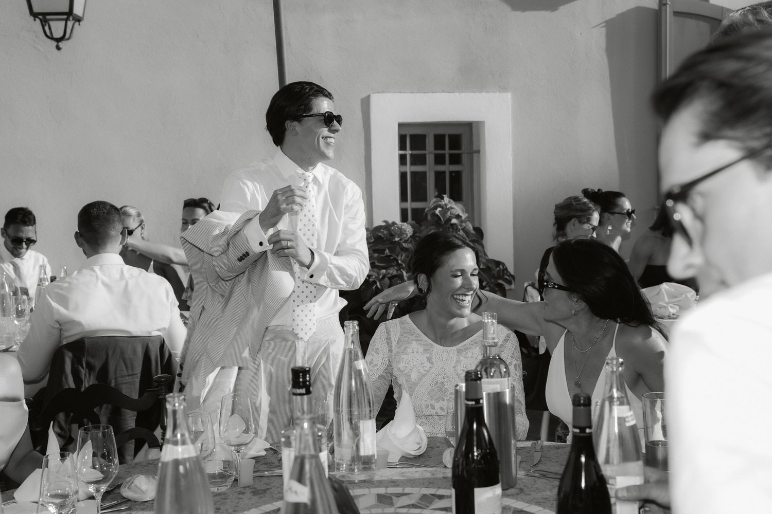 Black and white wedding reception photo with guests seated at tables, bride smiling, groom standing in suit and sunglasses.