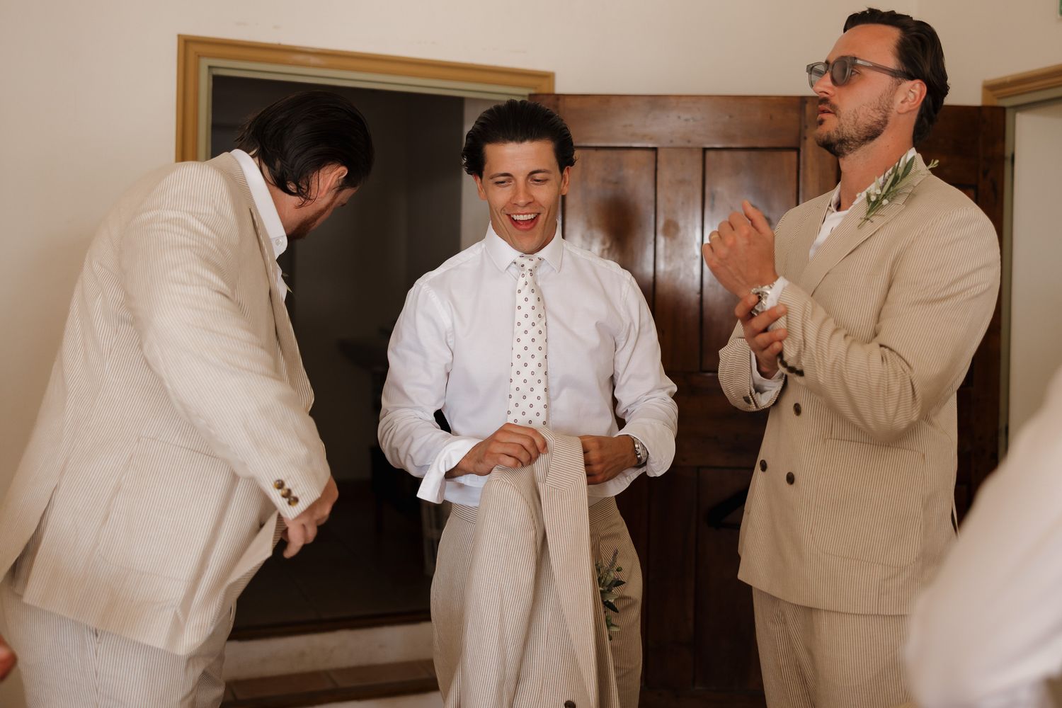 Three men in elegant suits laugh and celebrate together during a wedding preparation moment indoors.